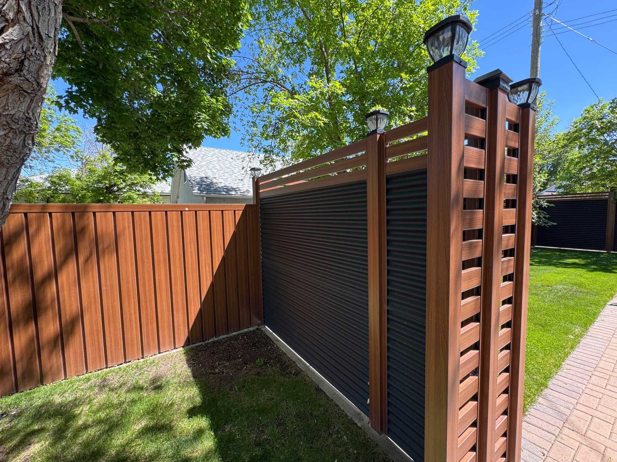 A wooden fence with vertical slats, green trees with full leaves, a blue sky, a house roof, and a sidewalk.