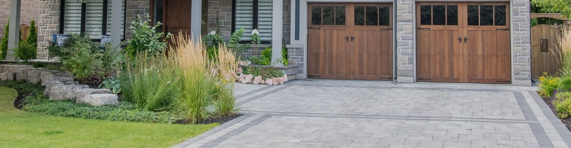 A modern house with a stone and wood exterior, featuring a garage with wooden doors and a landscaped front yard with grass, plants, and a stone-paved driveway.