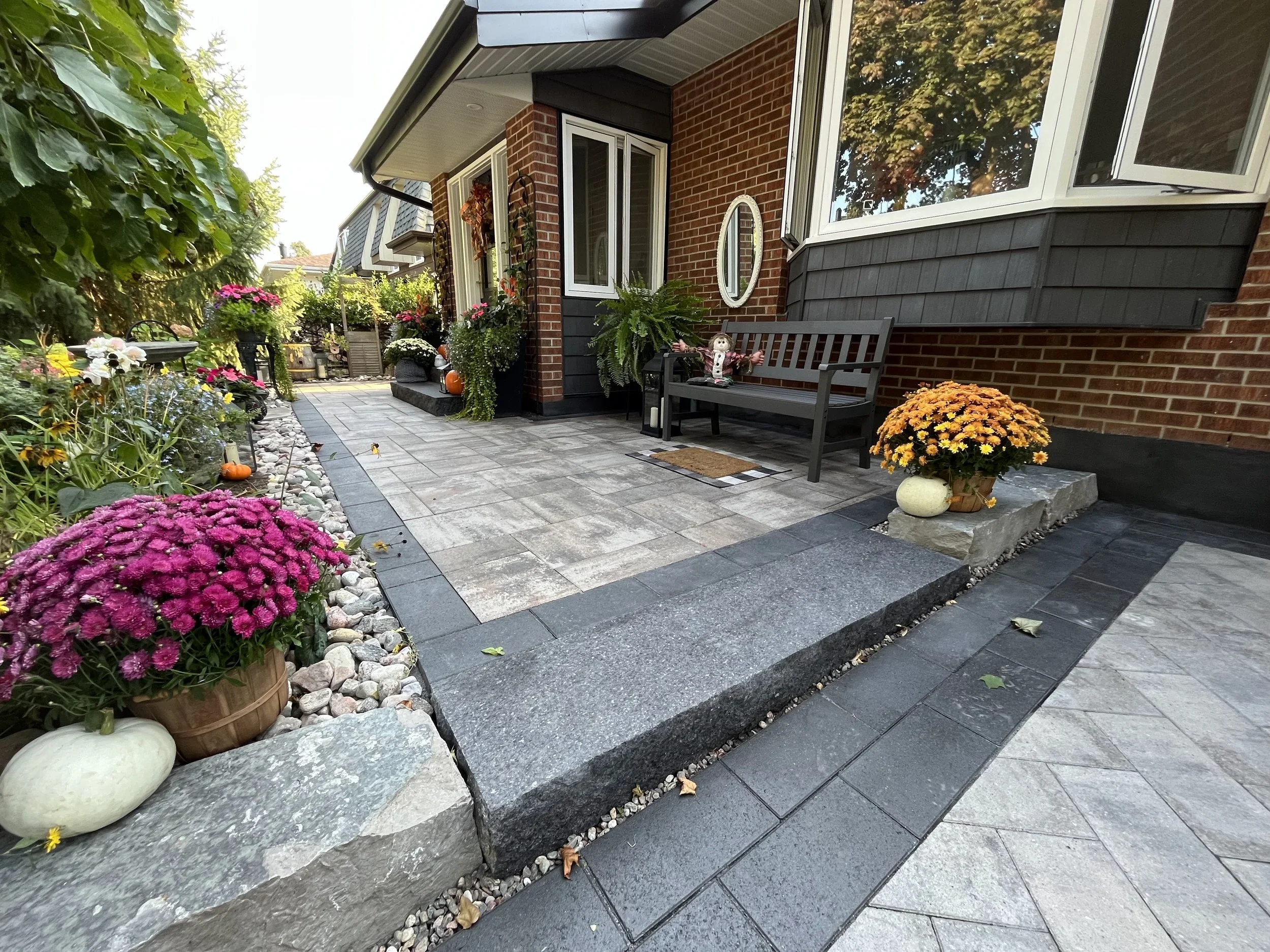 A front porch with potted plants, flowers, pumpkins, and decorative items, attached to a brick house with large windows and gray siding, surrounded by lush green trees.
