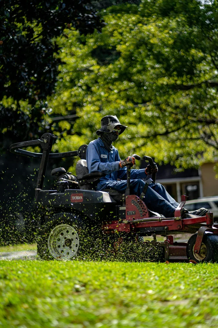 A person wearing a hat, mask, and gloves operating a riding lawn mower on a grassy lawn with trees in the background.