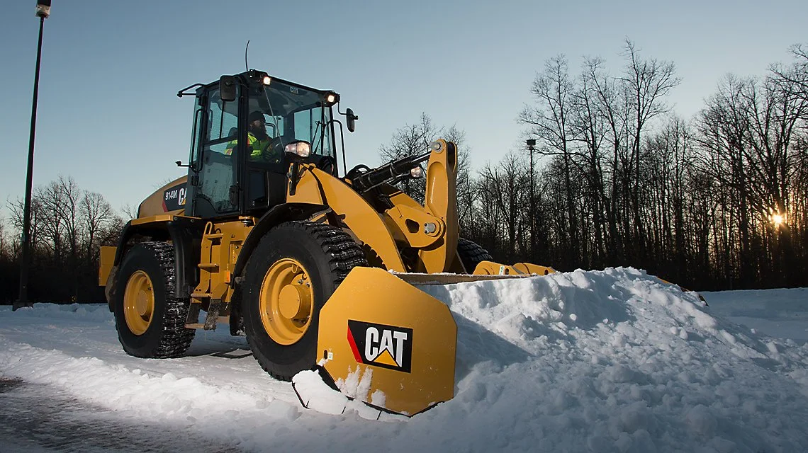 Yellow Caterpillar snowplow clearing snow on a snowy field with leafless trees and a clear sky at sunset.