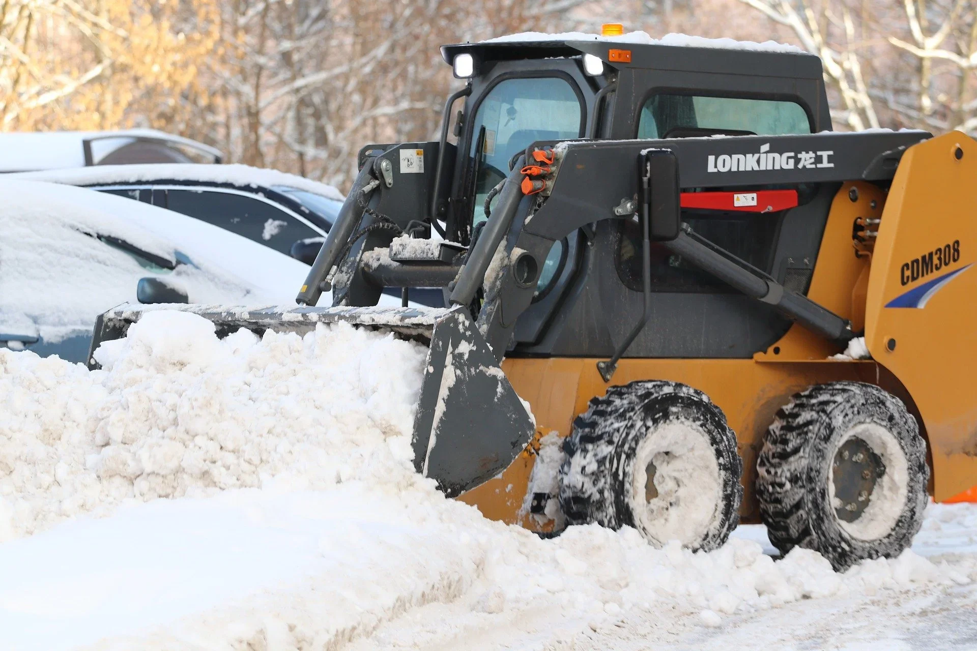 A yellow and black loader with the brand name "Lonking" and model number "CDM308" is clearing snow from a parking lot. Snow covers the ground and parked cars, with a background of trees and some snow on the branches.