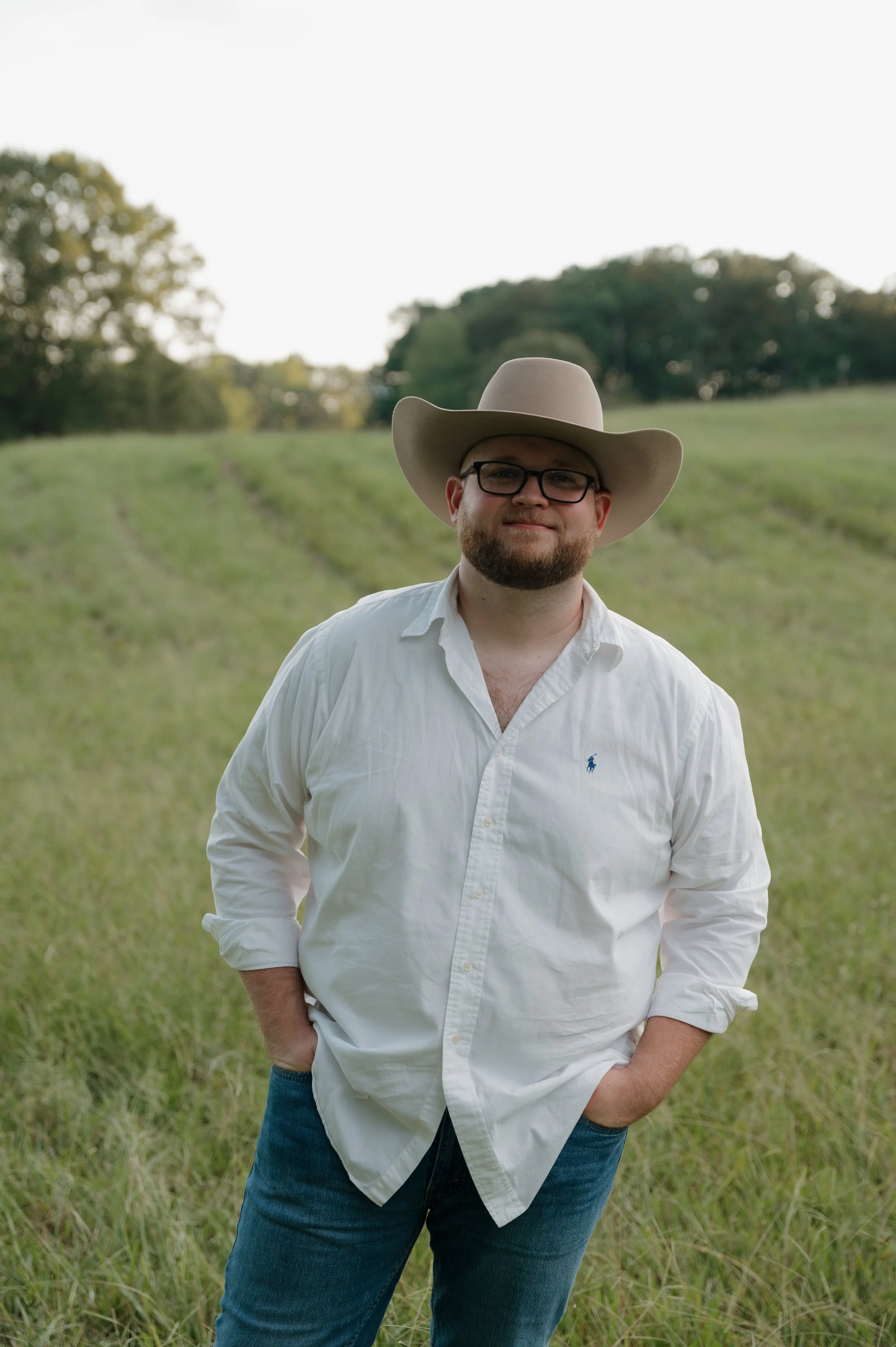 A man with glasses and a beard standing outdoors in a grassy field, wearing a white button-up shirt, dark jeans, and a large cowboy hat.