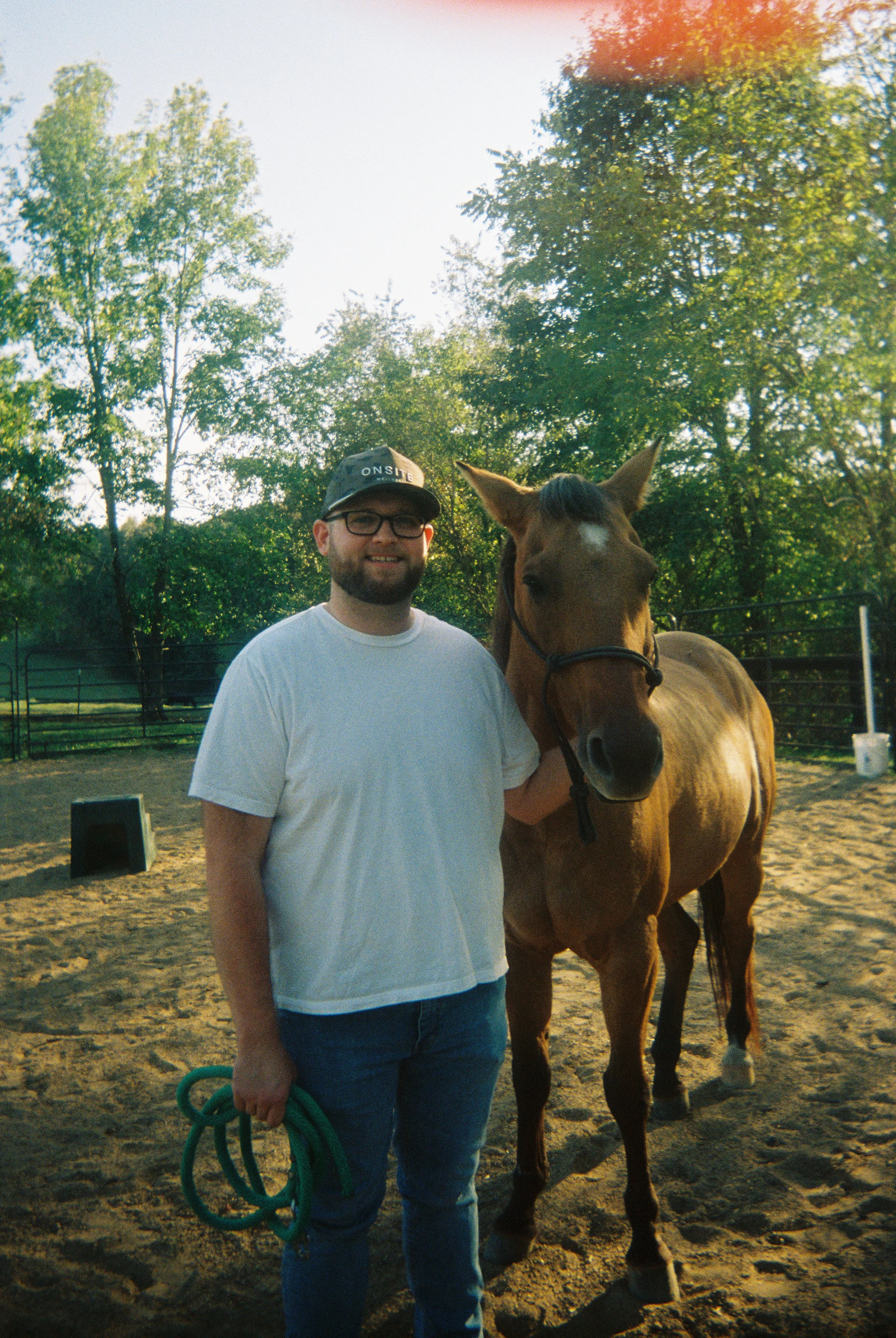 A man with glasses, a beard, and a black cap, wearing a white t-shirt and blue jeans, stands next to a brown horse with a white small mark on its forehead. The man is holding a green rope and is smiling. The background shows green trees and a fence, 