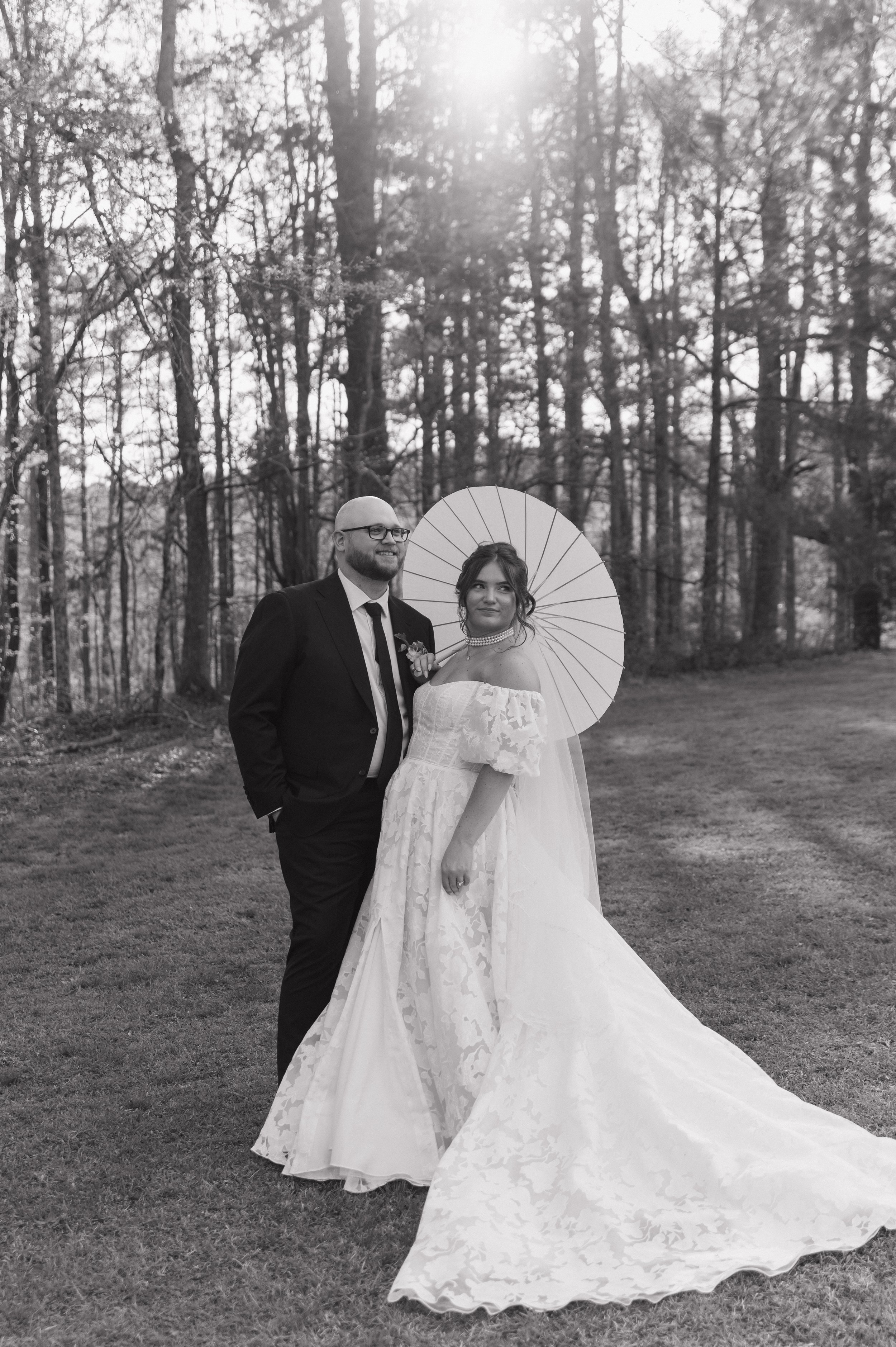 A couple in wedding attire standing outdoors in a forested area, with the woman holding a parasol and both smiling, black-and-white photo.