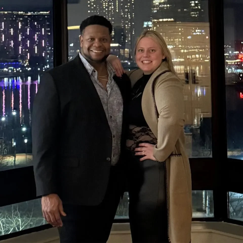 Two people, a man and a woman, smiling and posing together in front of a city skyline at night, with lights reflecting on the water.