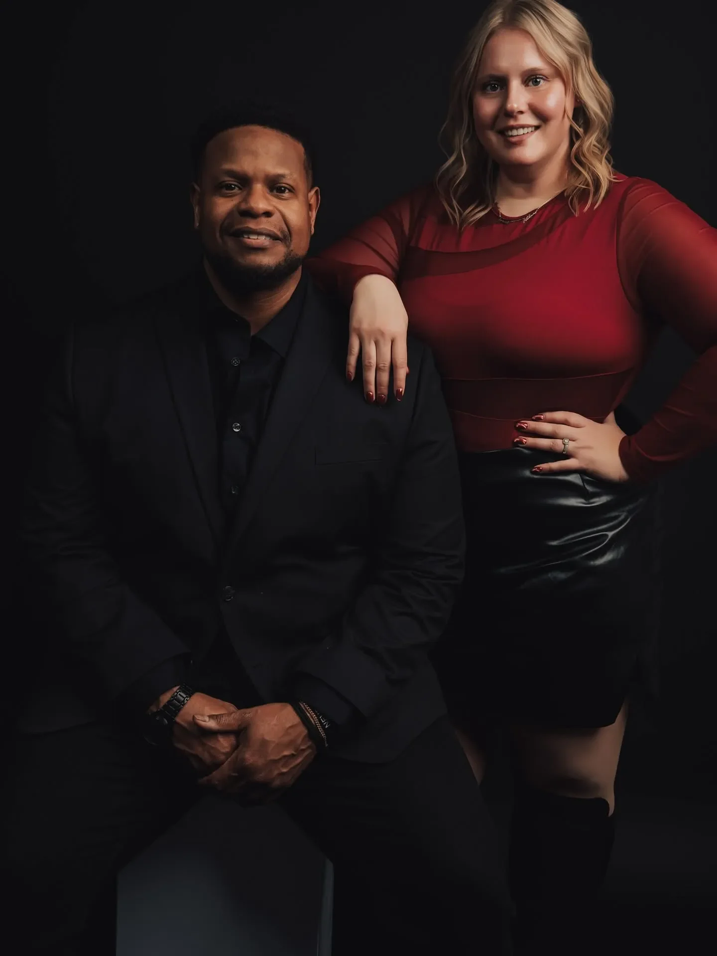A man and a woman posing together against a dark background. The man is seated, wearing a black suit, and has a dark complexion. The woman, standing next to him with her hand resting on his shoulder, has blonde wavy hair, fair skin, and is wearing a red, long-sleeved top and a black skirt.