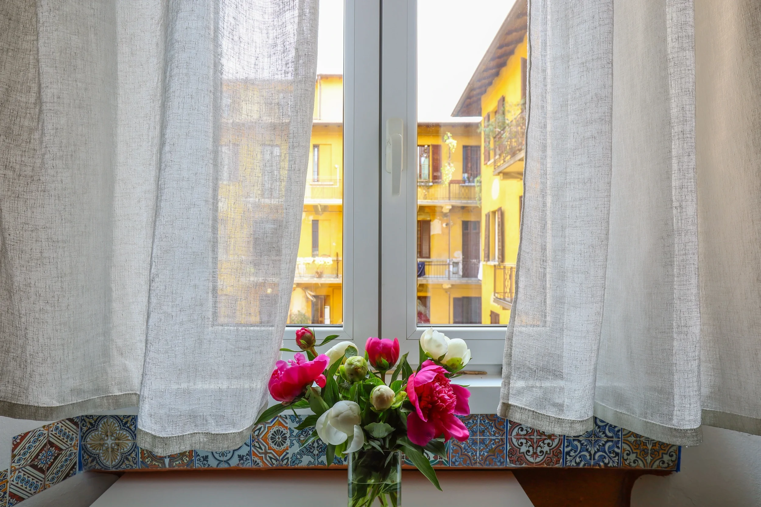 Peonies on the vintage-tiled sill, linen curtains and the yellow courtyard beyond