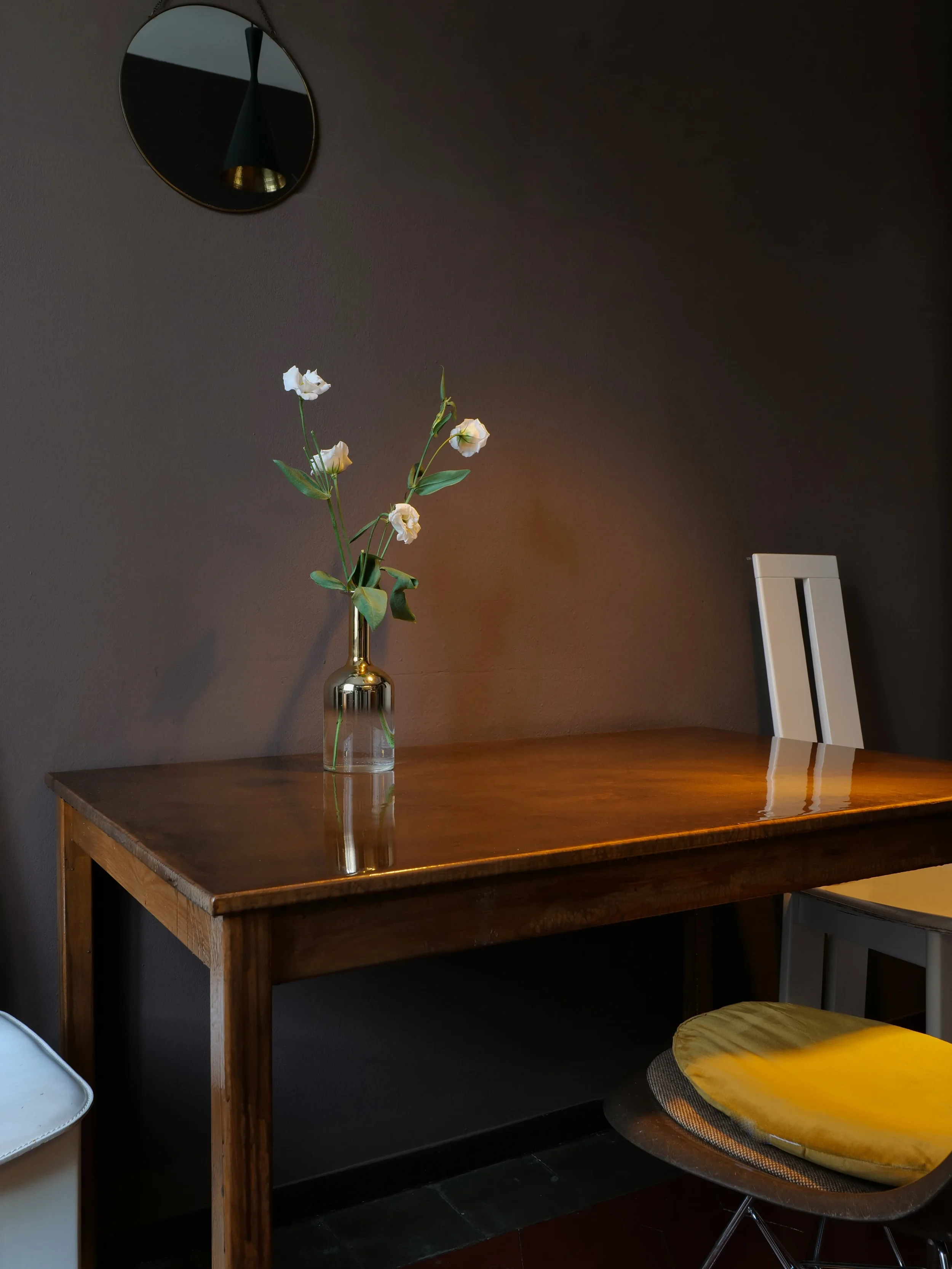 Dining area with dark brown wall, wooden table, round mirror and floral arrangement, House of Frames Milan