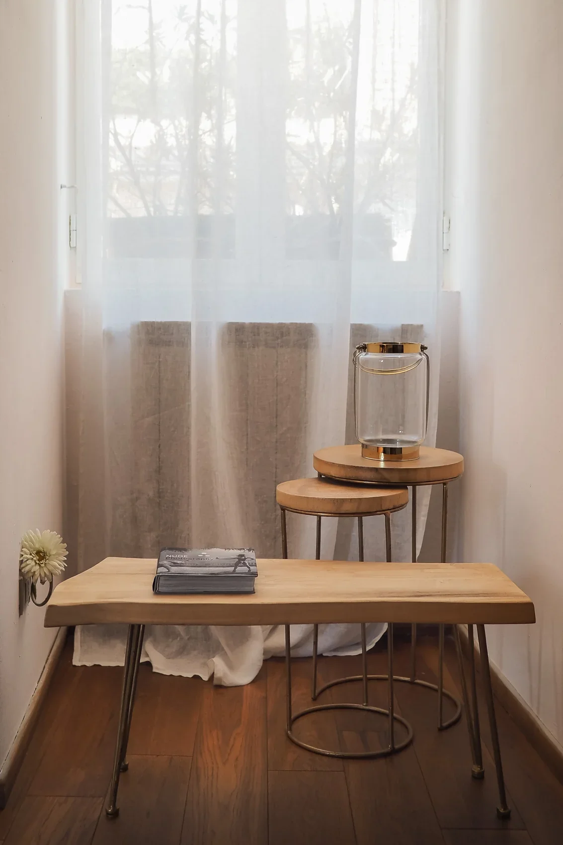 Wooden bench and nesting side tables by the window, with linen curtains and a glass vessel