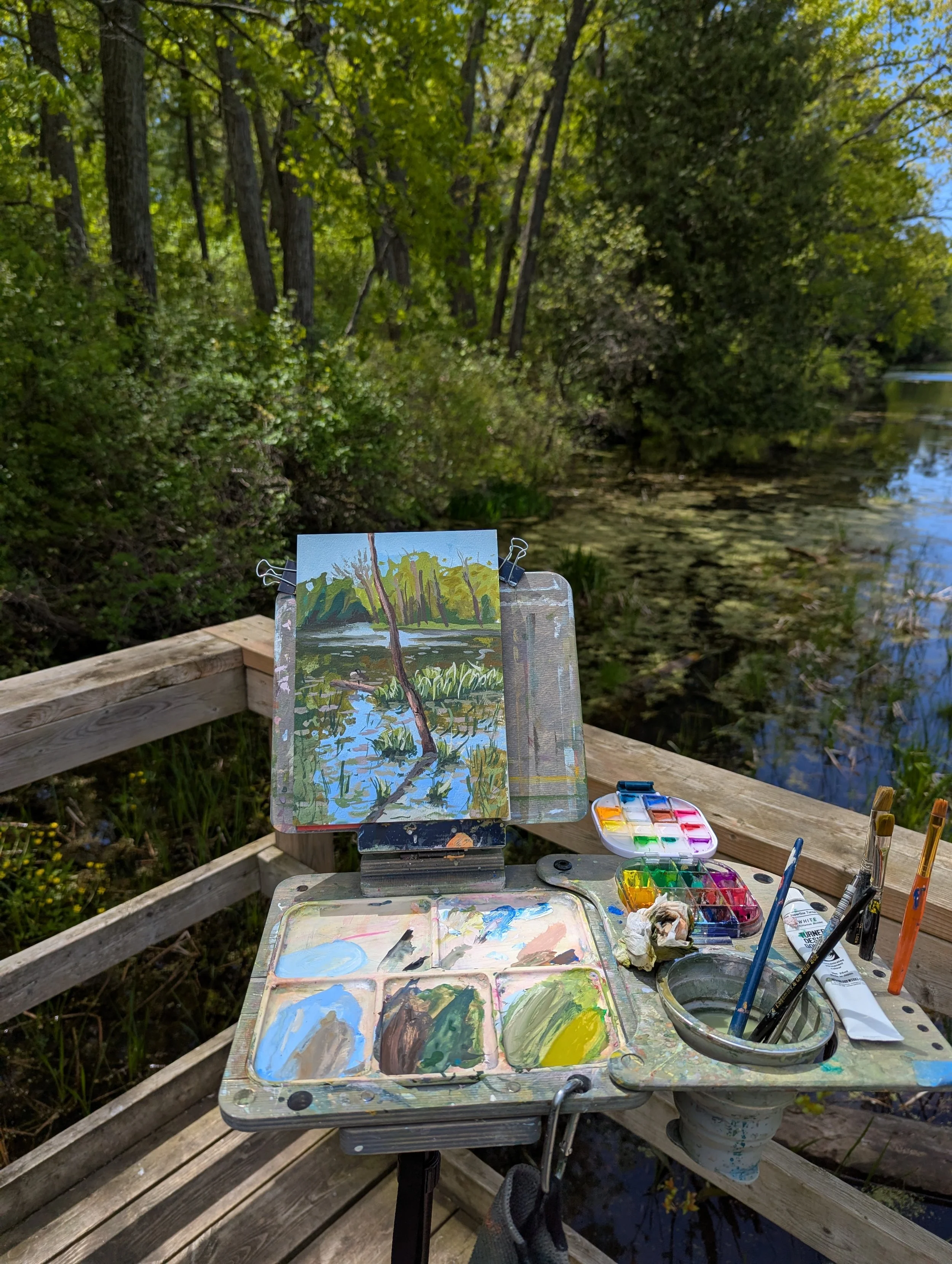 An outdoor painting setup with a small canvas on an easel depicting a nature scene with trees and water, surrounded by painting supplies including watercolors, brushes, and a palette, on a wooden deck near a body of water with lush green trees and a 