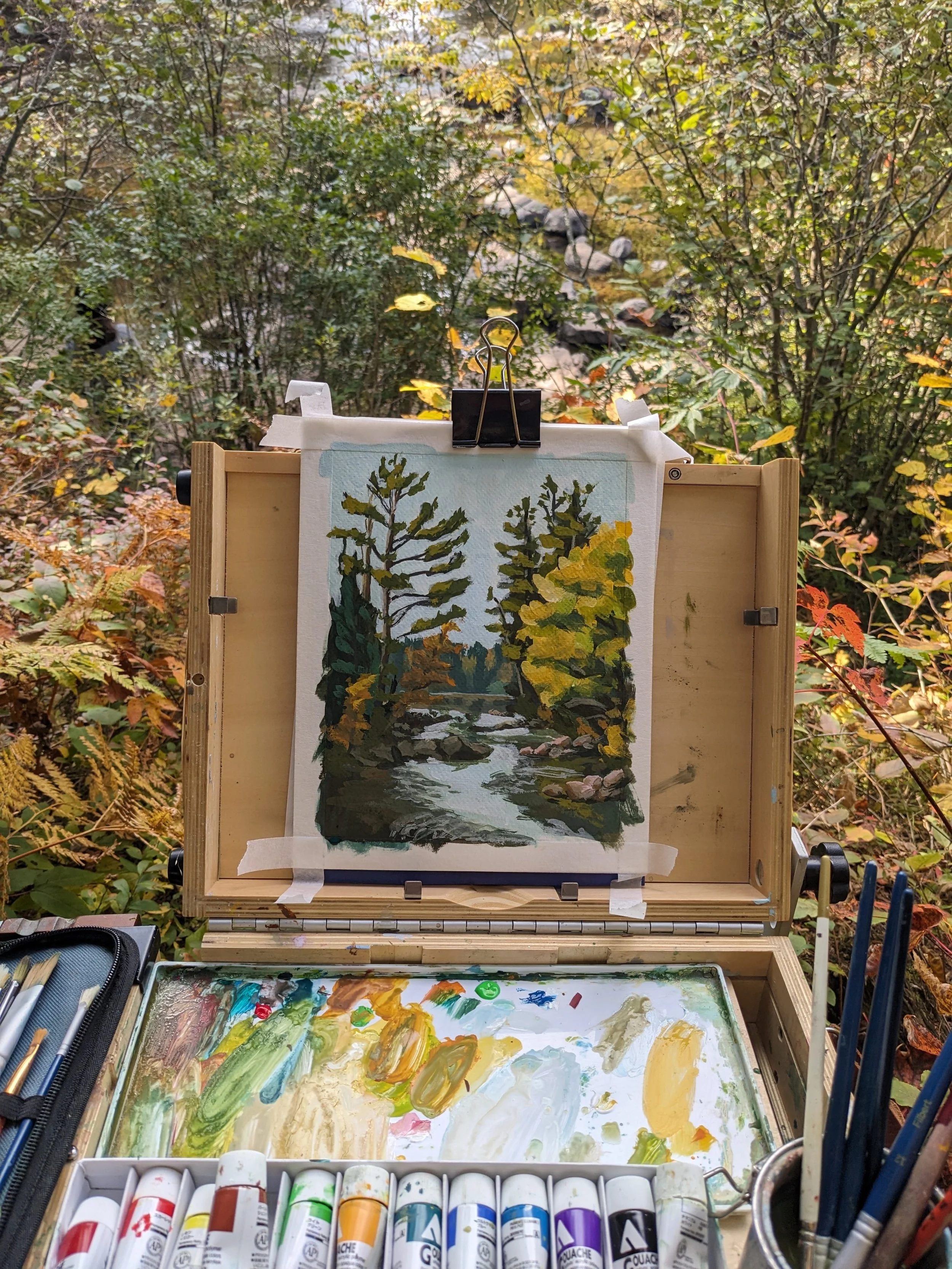 An outdoor plein air painting setup with a landscape watercolor painting of trees and a river, surrounded by art supplies including paint tubes, brushes, and a palette.