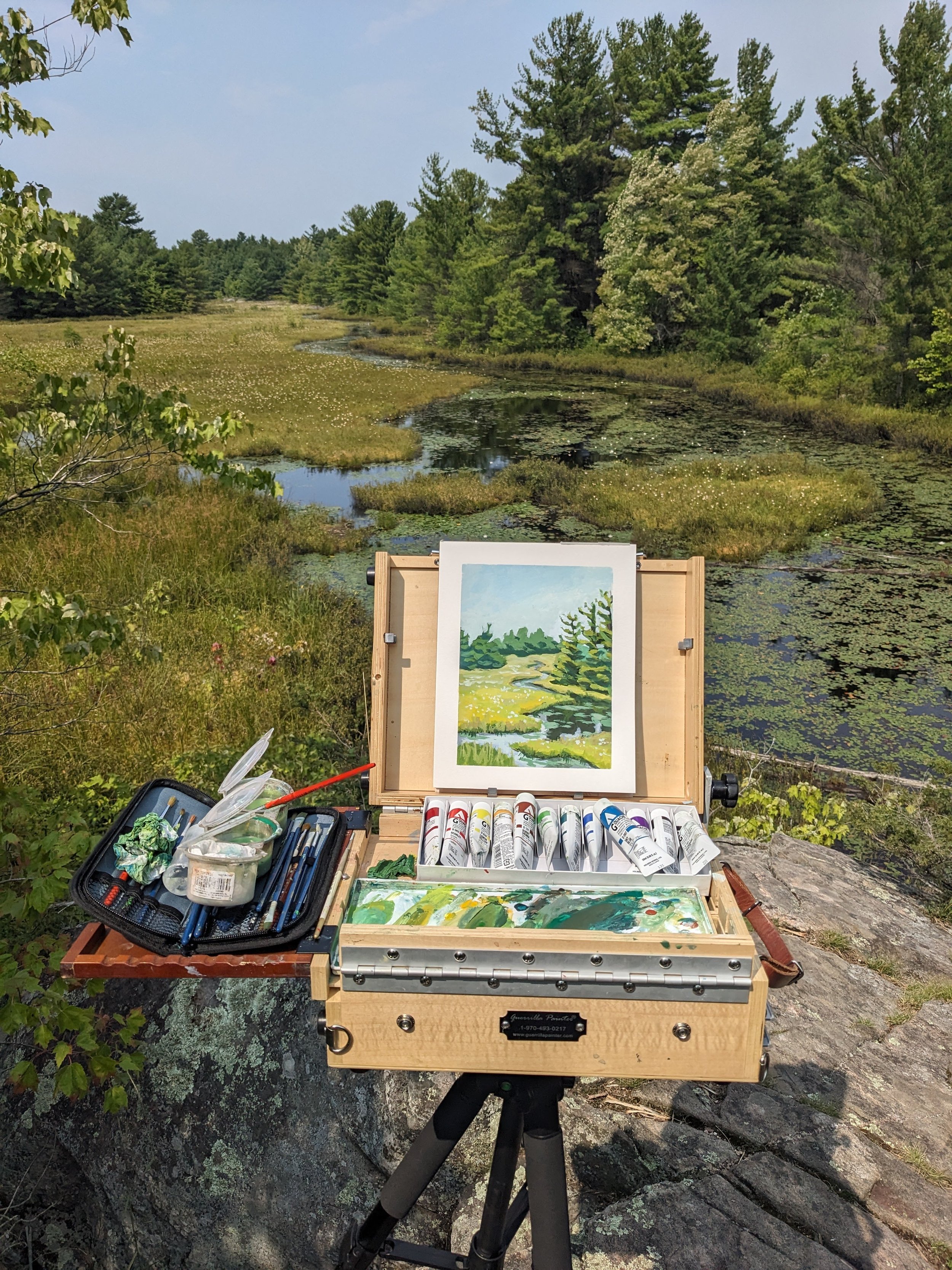 A painting easel and art supplies set up outdoors on a rocky surface by a pond surrounded by trees, with a scenic landscape painting of a pond, trees, and sky on the easel.