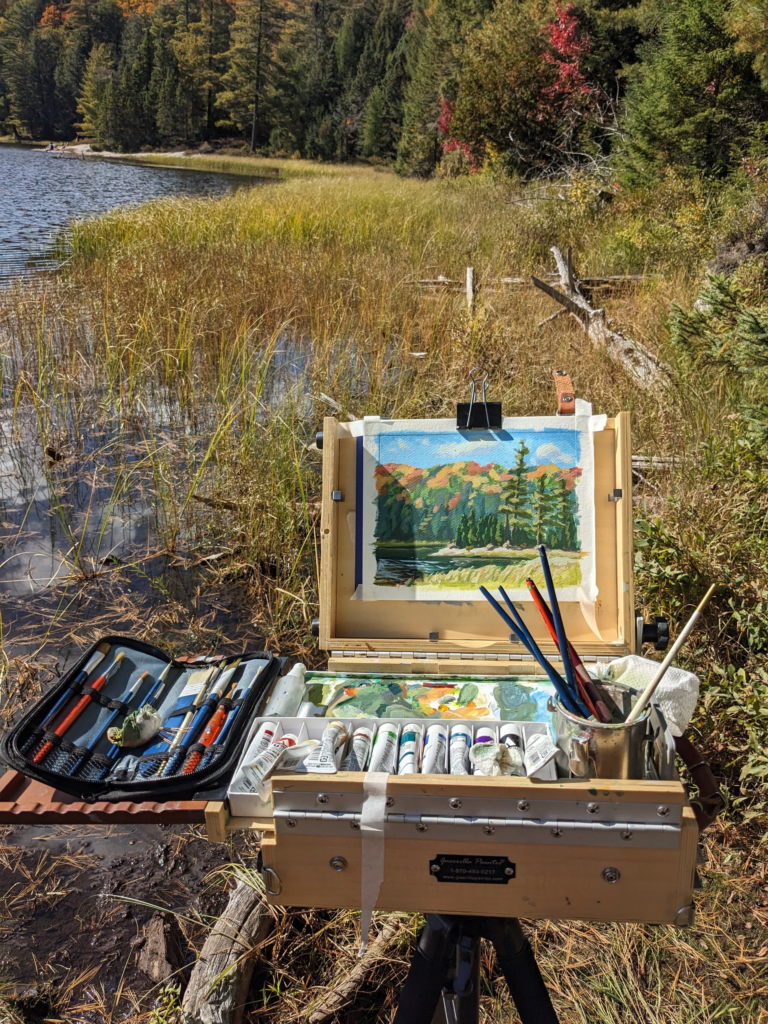 A painting setup outdoors near a lake with trees, including a watercolor landscape of trees and mountains on an easel, surrounded by paint tubes, brushes, and art supplies.