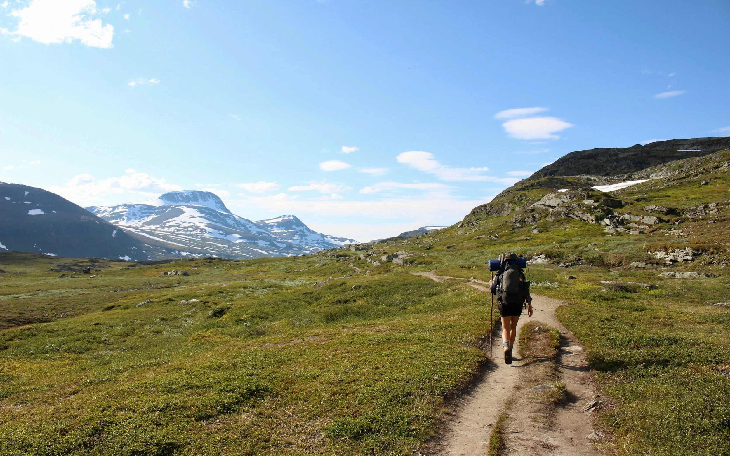 A person hiking on a trail through a mountainous landscape with snow-capped peaks, green hills, and a blue sky with scattered clouds.
