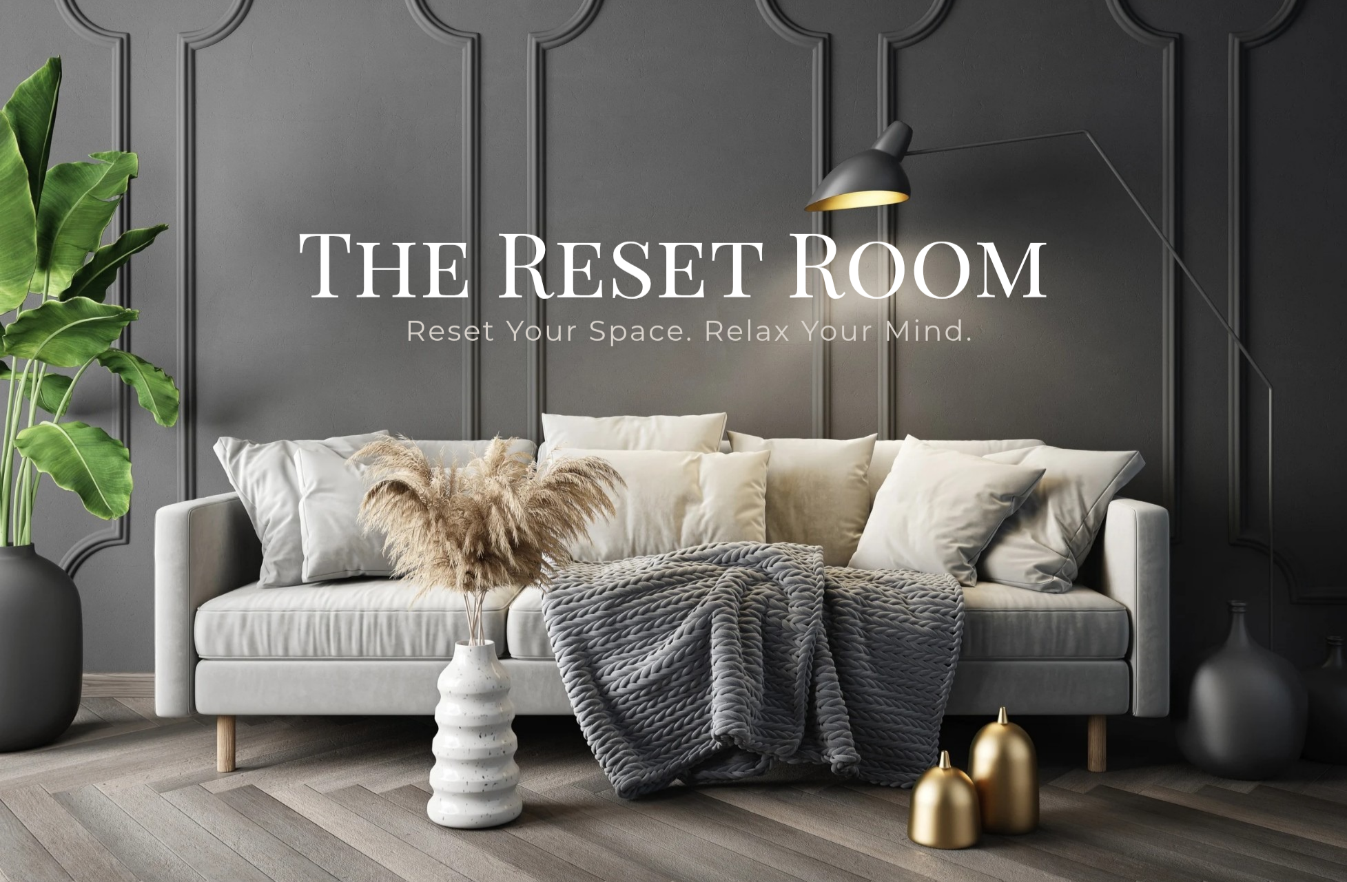 A cozy living room with a white sofa, beige cushions, a dark gray knitted throw blanket, and a white vase with pampas grass on the floor. There is a large green potted plant on the left and dark gray decorative wall paneling behind the sofa. Two gold decorative objects are on the floor, and a black wall sconce lamp is mounted on the wall.
