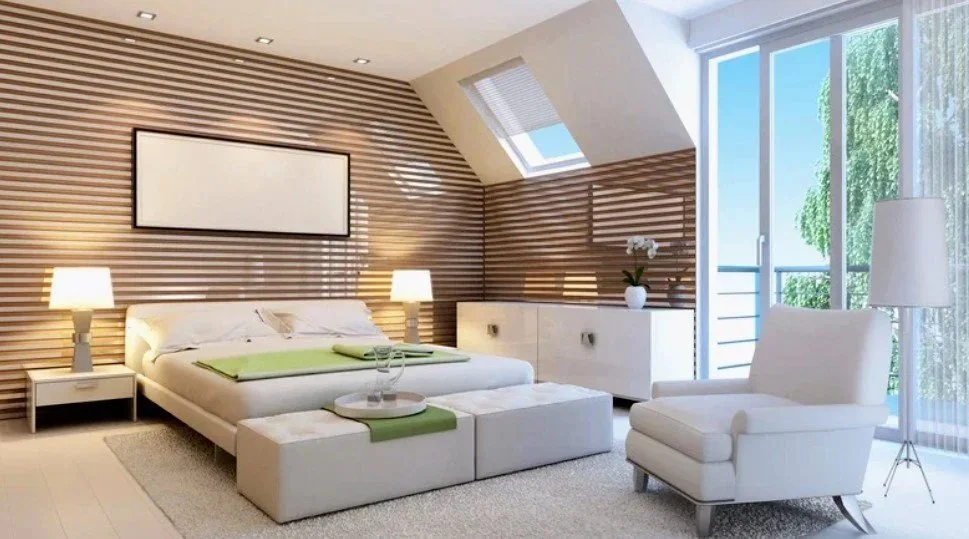 Modern hotel room with a bed, two nightstands with lamps, an armchair, a cabinet with a potted plant, a framed picture on a wooden wall, and large windows with a balcony overlooking trees.