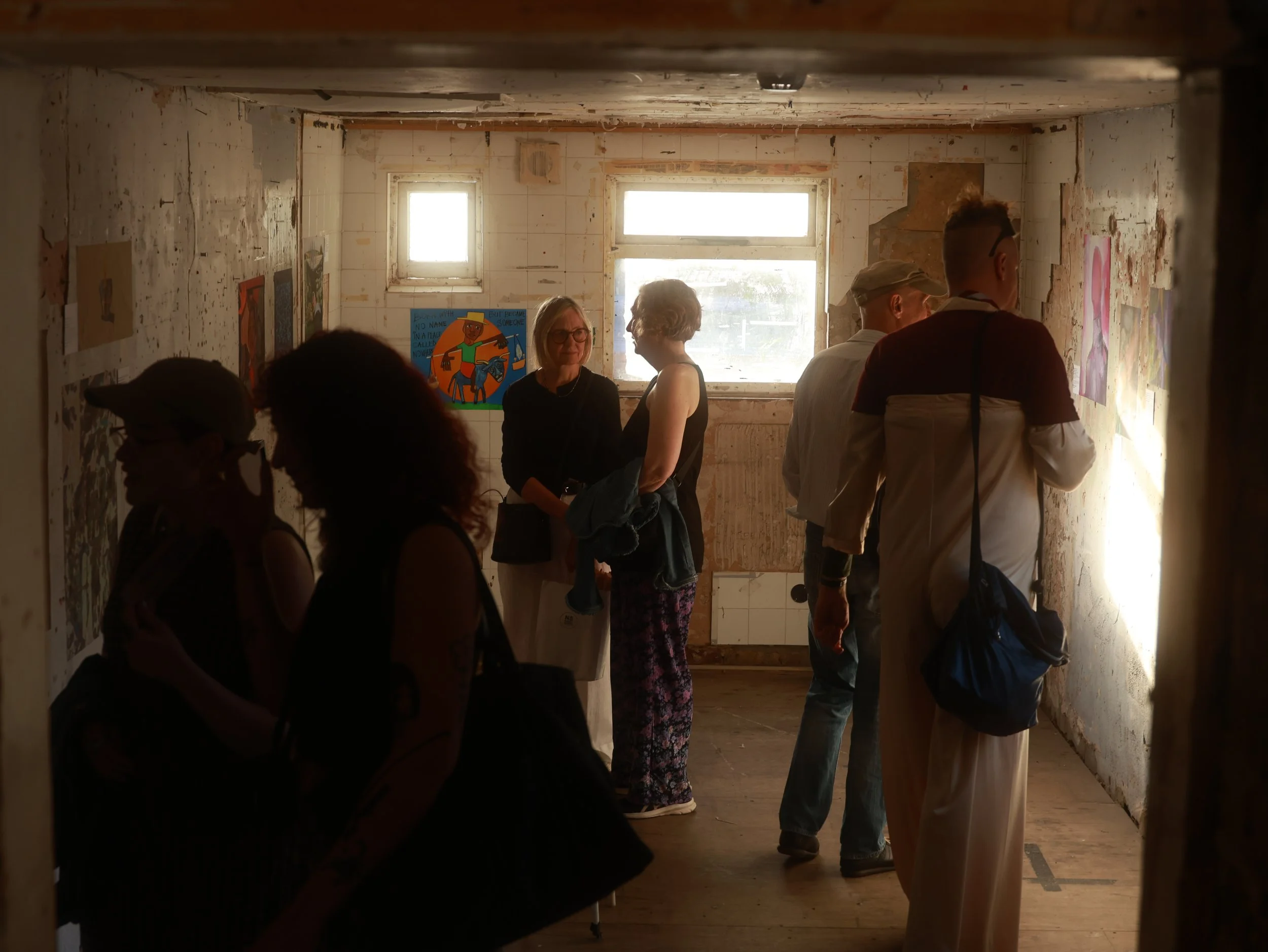 People standing and talking inside an unfinished room with peeling paint and worn walls, illuminated by natural light from windows.