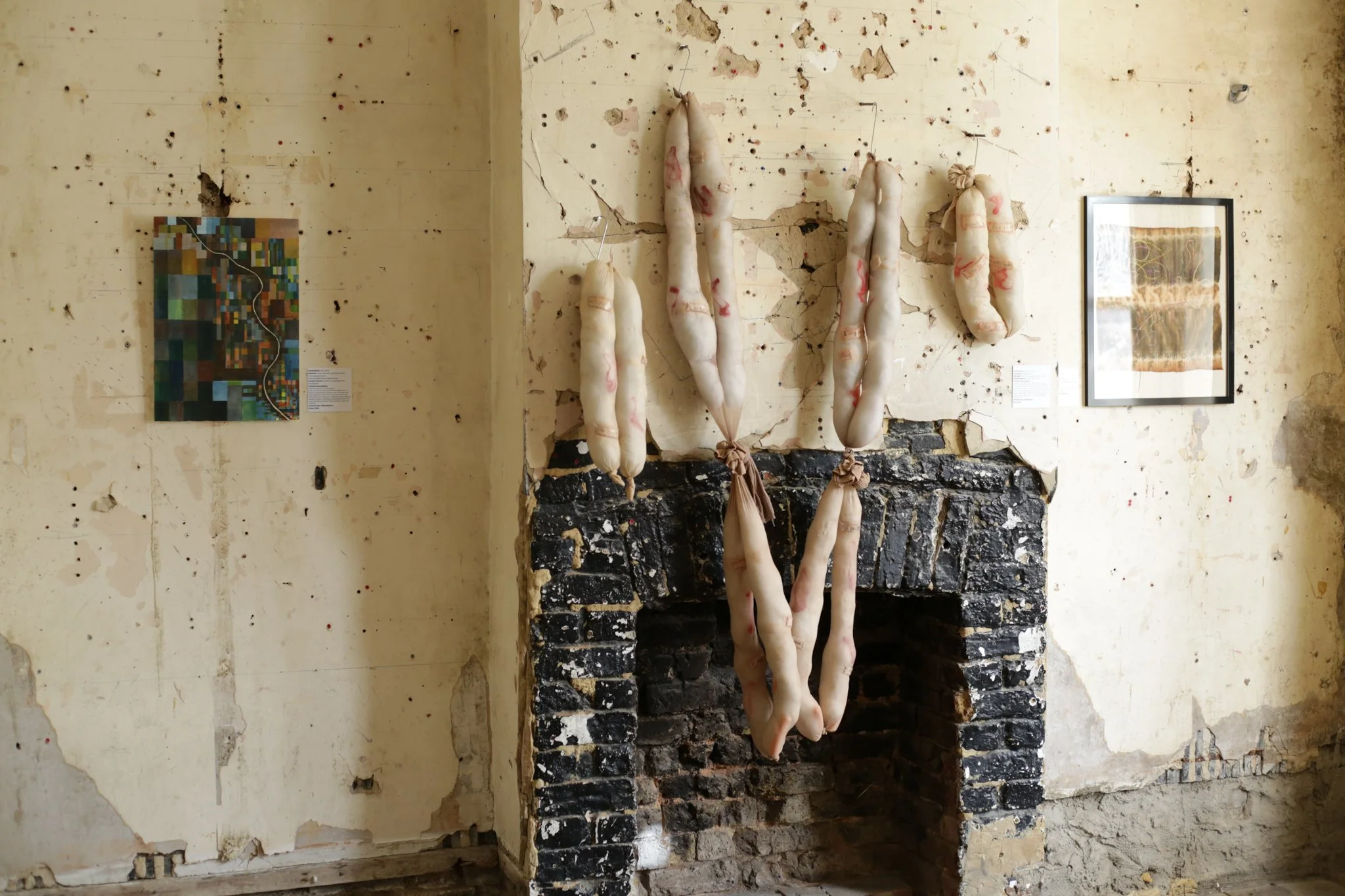 The image shows an art installation with seven fabric objects hanging vertically on a distressed, cracked wall, with two framed pieces of art on either side of the hanging pieces.