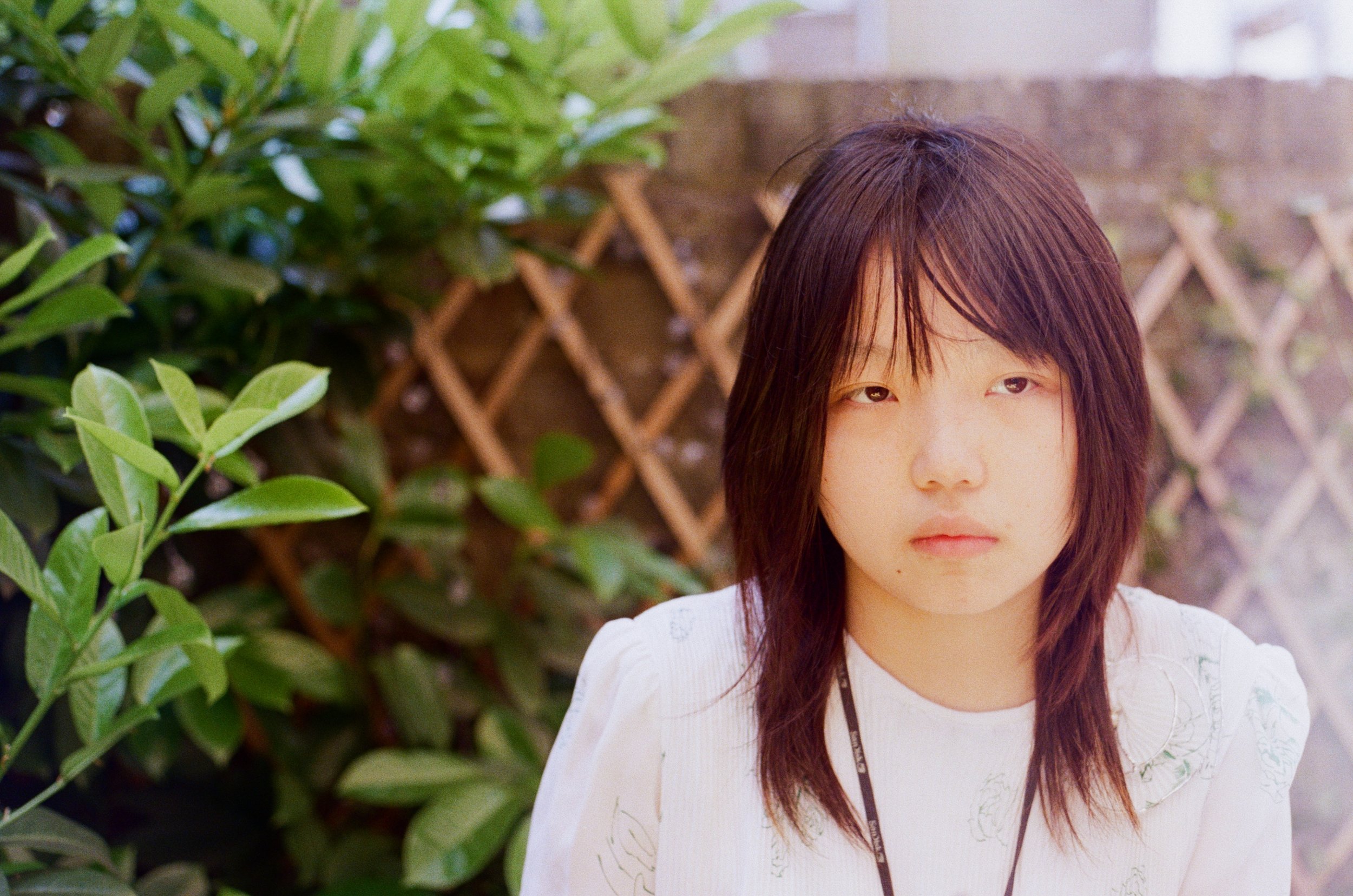 Young woman with shoulder-length dark hair, wearing a white shirt, standing outdoors near green leafy plants and lattice fencing.