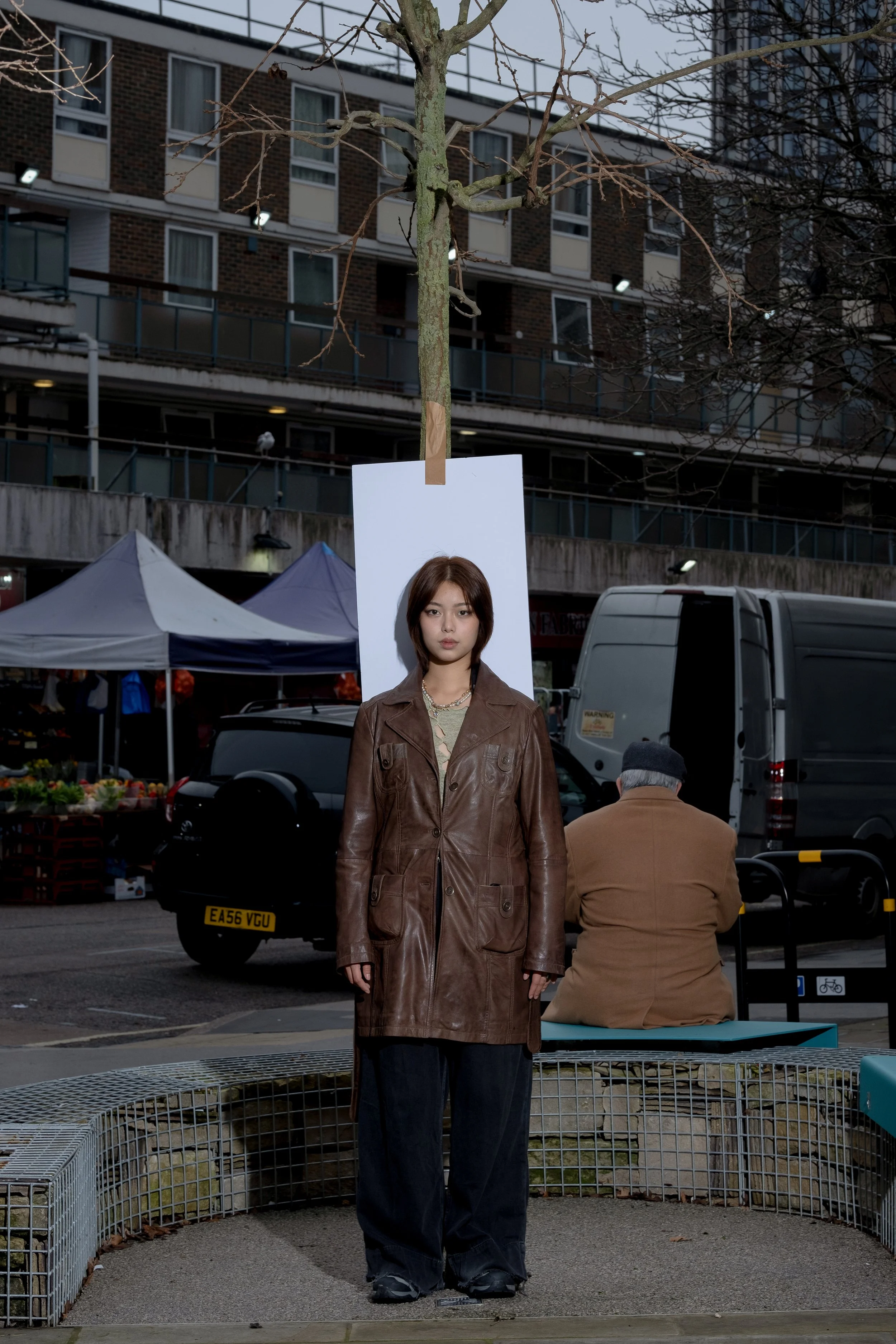 A woman standing in front of a white board with a leafless tree behind her in an urban market setting with tents and a building in the background.