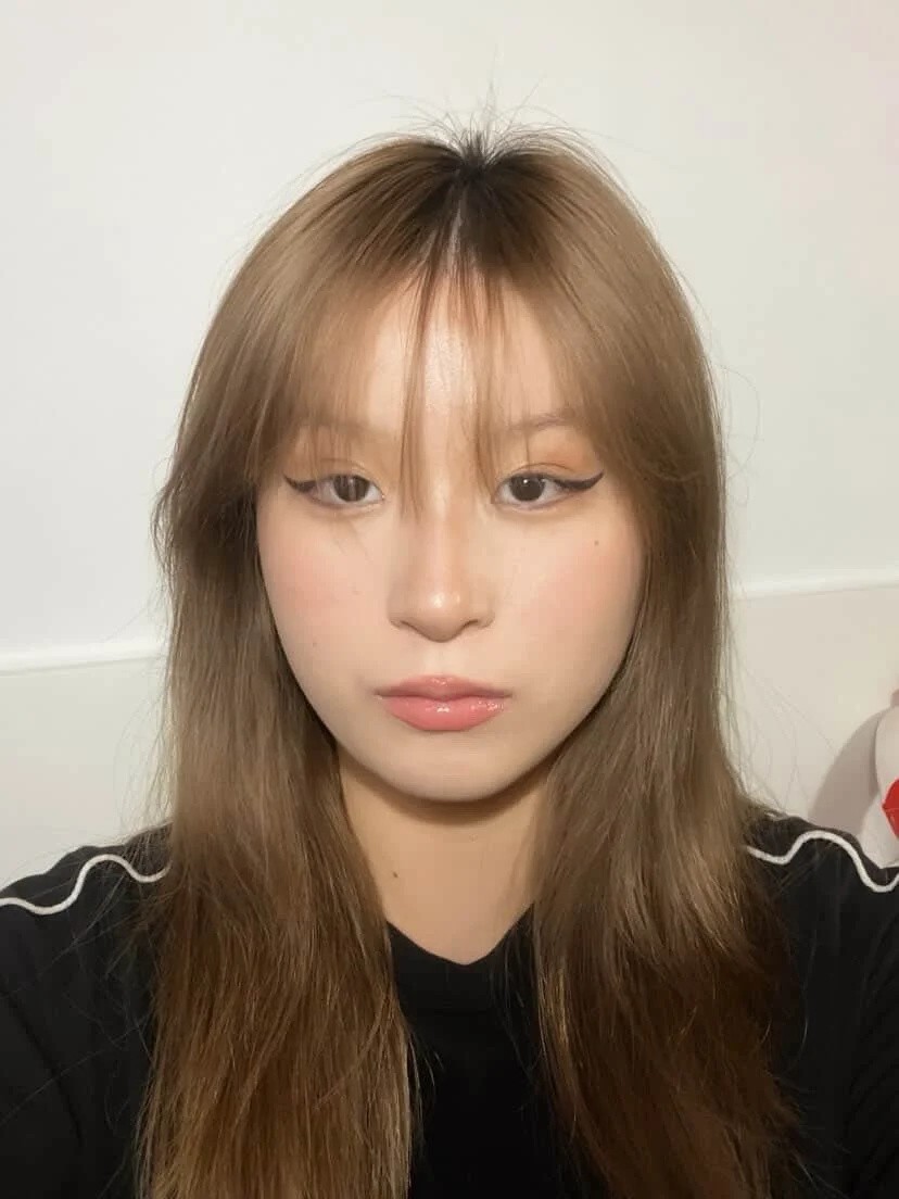 Close-up photo of a woman with light brown, slightly wavy hair, wearing makeup with winged eyeliner and natural lipstick, looking at the camera against a plain white wall.