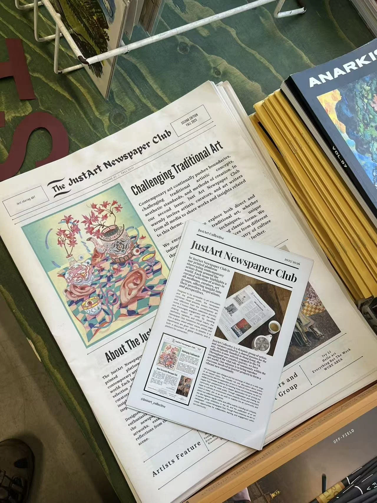 A newspaper titled "The JustArt Newspaper Club" with an article about challenging traditional art, a colorful art illustration, and a small flyer with similar information. The newspaper is on a table with a stack of art books nearby.