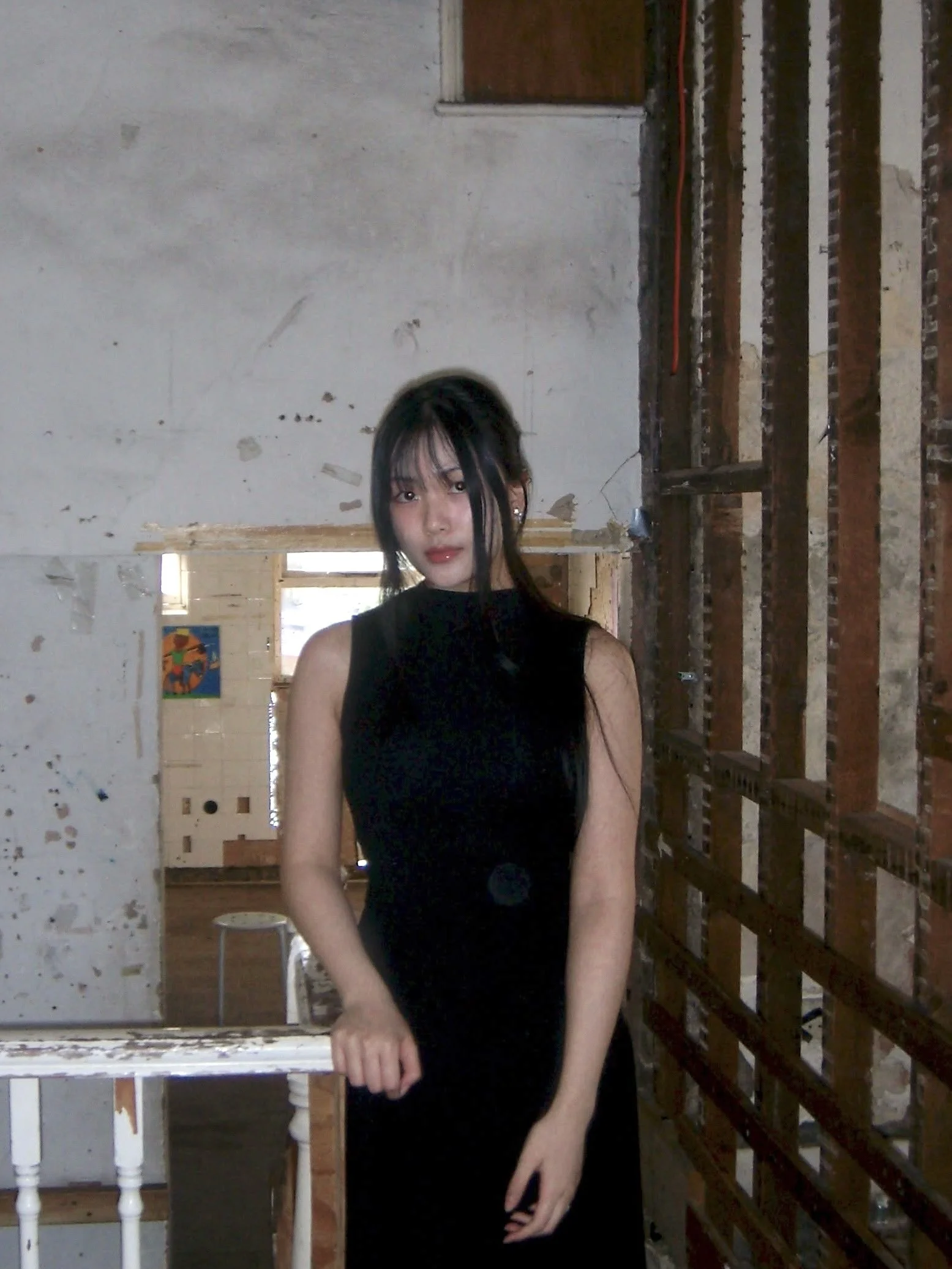 A young woman with black hair and makeup, wearing a sleeveless black dress, standing in an unfinished room with exposed brick wall and construction materials.