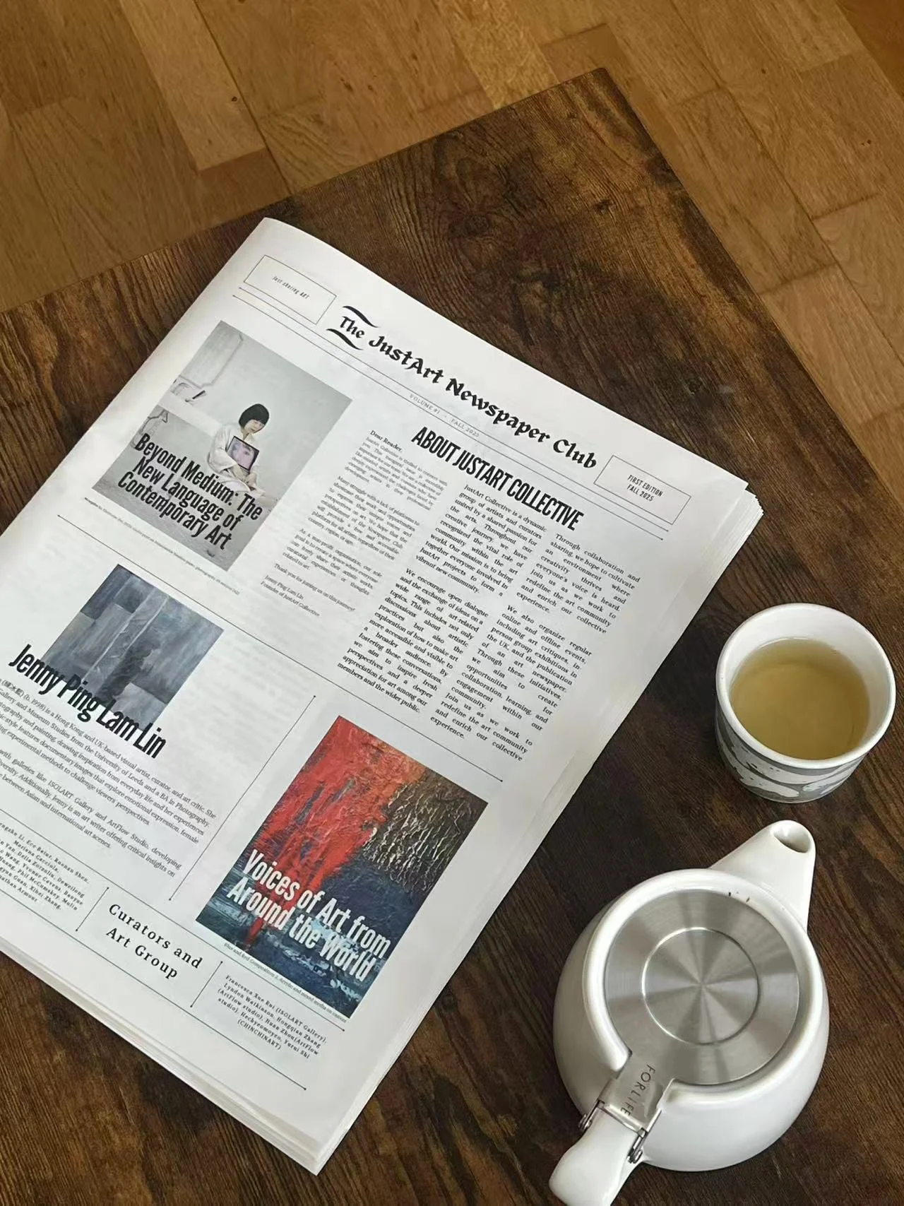 A newspaper titled 'The JusstArt Newspaper Club' on a wooden table alongside a cup of yellowish-green tea, a white teapot, and a white teapot lid.