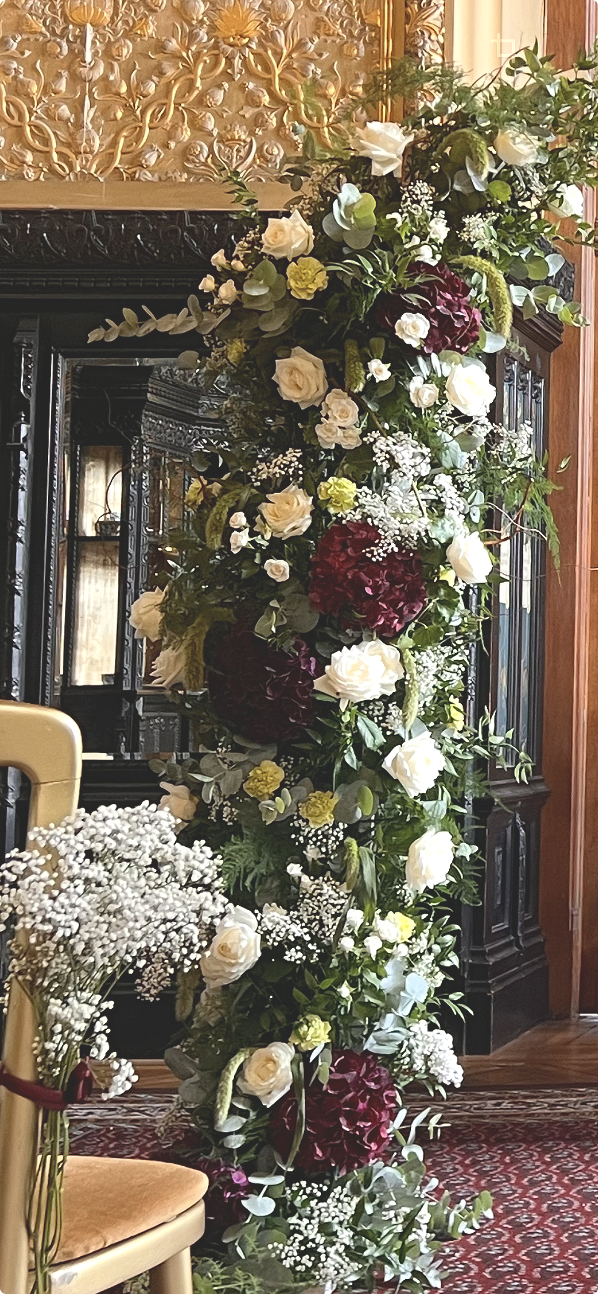 A large floral arrangement with white roses, burgundy flowers, and greenery, set against ornate black furniture and gold decorative elements in a decorated indoor setting.