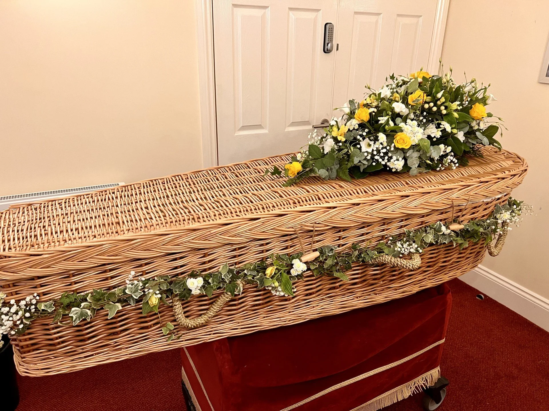 A wicker casket decorated with a large bouquet of yellow and white flowers and green foliage, sitting on a red velvet stand indoors.