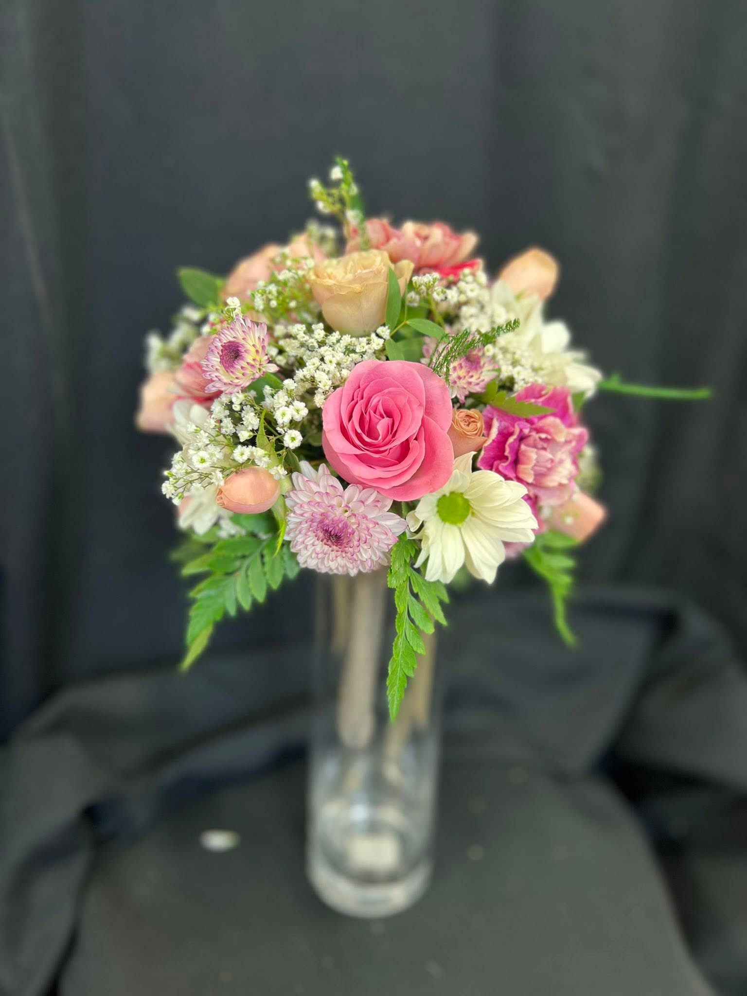 A bouquet of pink, white, and pastel flowers in a clear glass vase on a dark background.