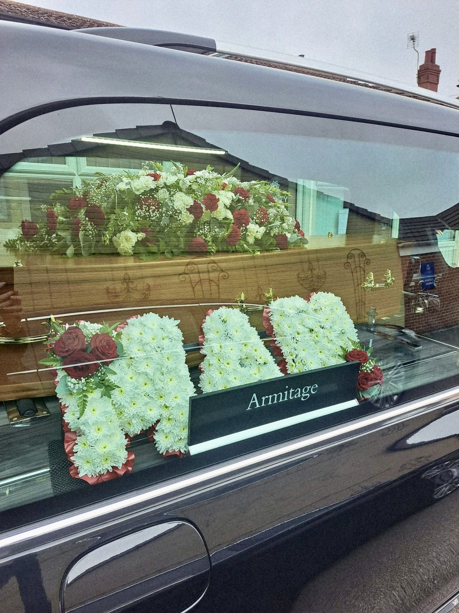 A hearse decorated with flowers, including white and red roses and other floral arrangements, parked on a street. The vehicle has a black exterior and the nameplate reads "Armitage."