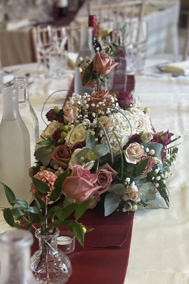A floral centerpiece with pink, peach, white, and burgundy roses, baby's breath, and greenery on a table with white tablecloth and wine bottles.
