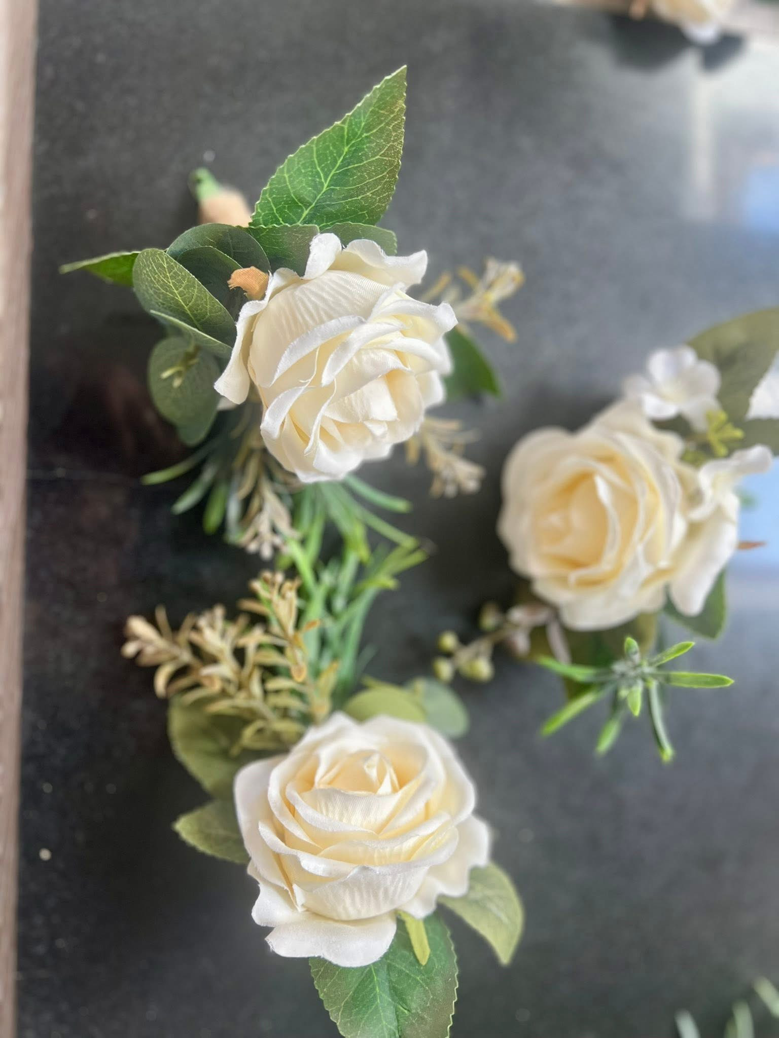 A small arrangement of white roses, green leaves, and sprigs of small white flowers on a dark surface.