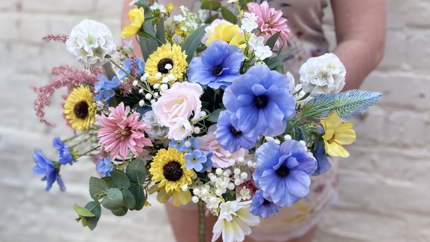 A person holding a colorful bouquet of various flowers, including blue, yellow, pink, white, and purple blossoms, in front of a light-colored brick wall.