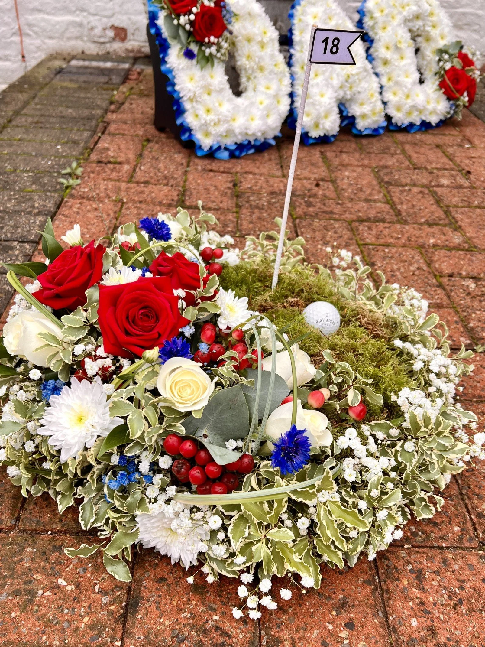 Floral arrangement on brick surface commemorates a memorial, with a small flag marked '18' and a golf ball among the flowers.