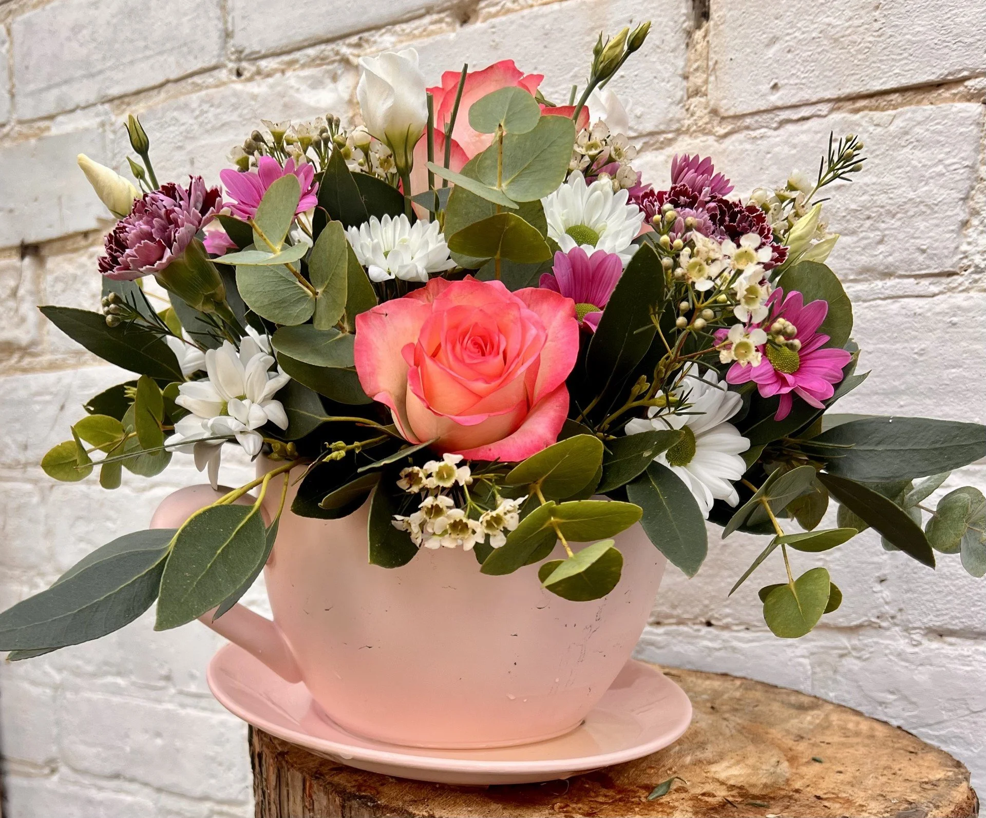 Pink flower arrangement in a pink ceramic teapot with eucalyptus leaves, white daisies, pink chrysanthemums, and other small flowers, set against a white brick wall background.