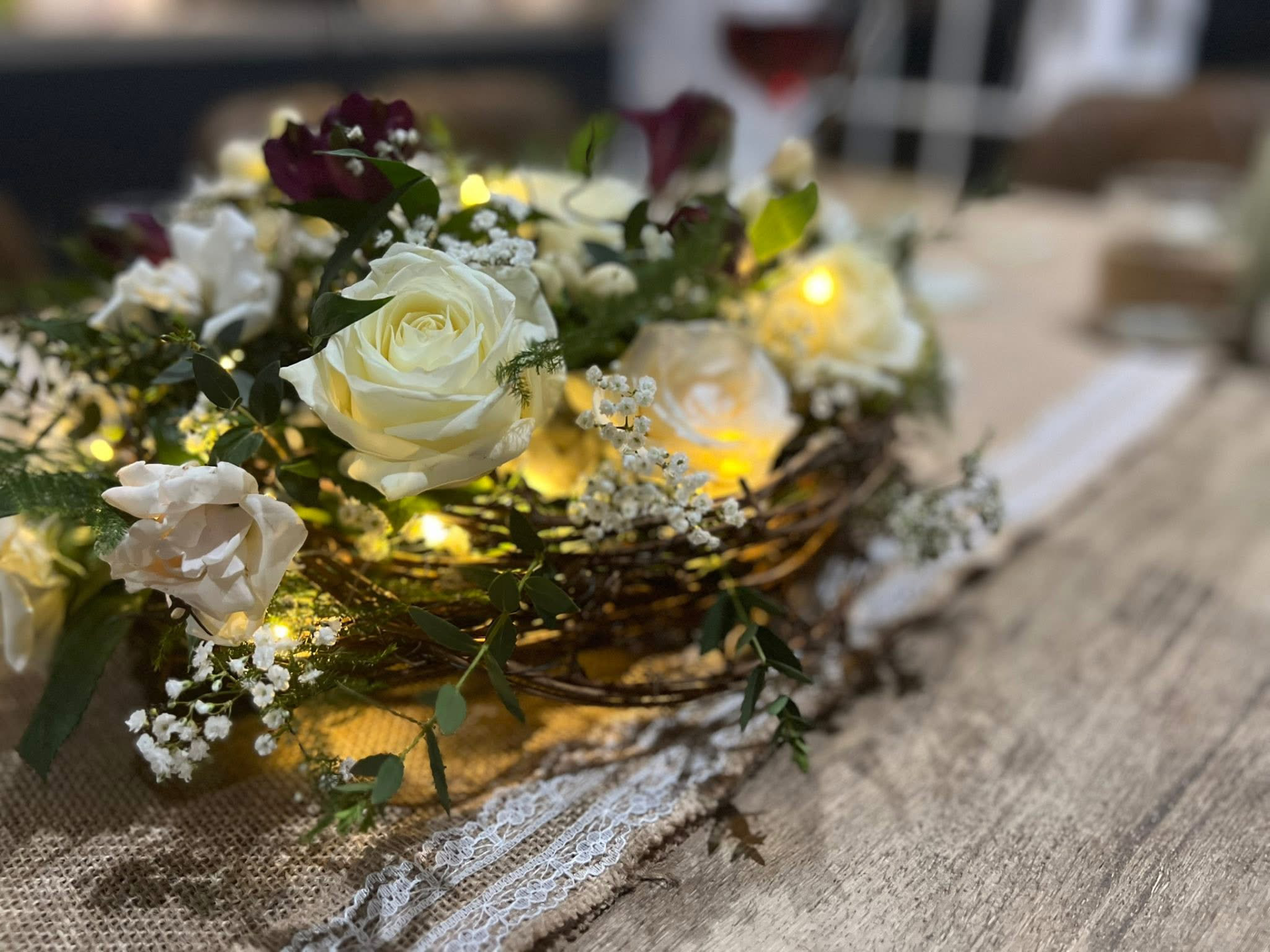 A floral arrangement with white roses, small white flowers, and green leaves in a basket, illuminated with warm yellow lights on a lace cloth on a table.