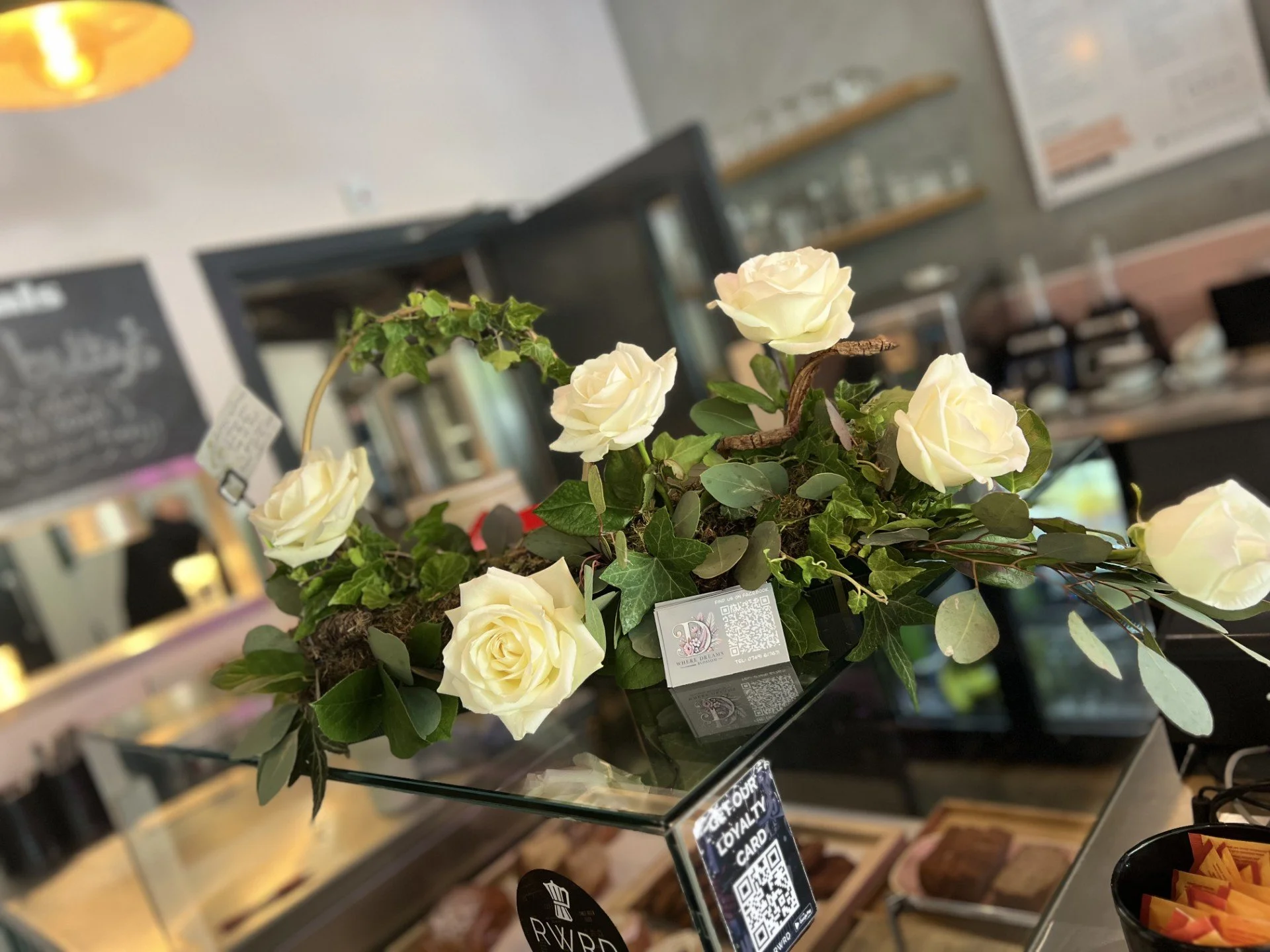 A bouquet of white roses and green foliage on a glass display counter in a cafe or restaurant.
