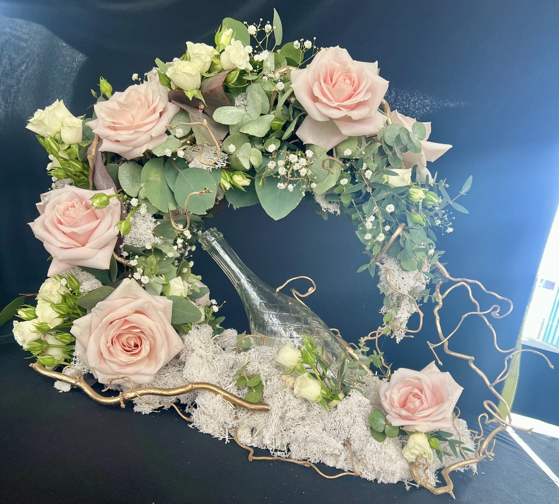 Arrangement of light pink roses, small white flowers, greenery, and decorative gold twigs in a glass vase on a black table against a blue background.