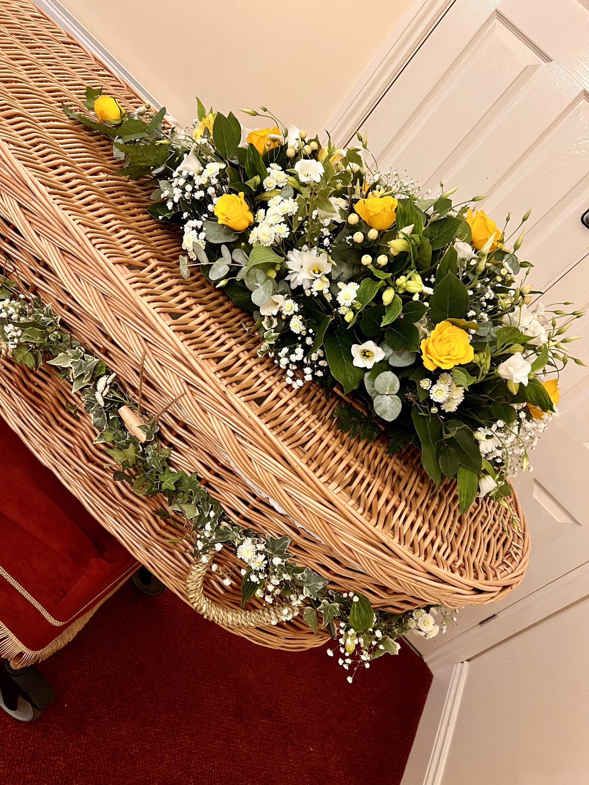 A wicker basket with a floral arrangement of yellow roses, white daisies, and green foliage, decorated with ivy, placed in a room with beige wall and white door.