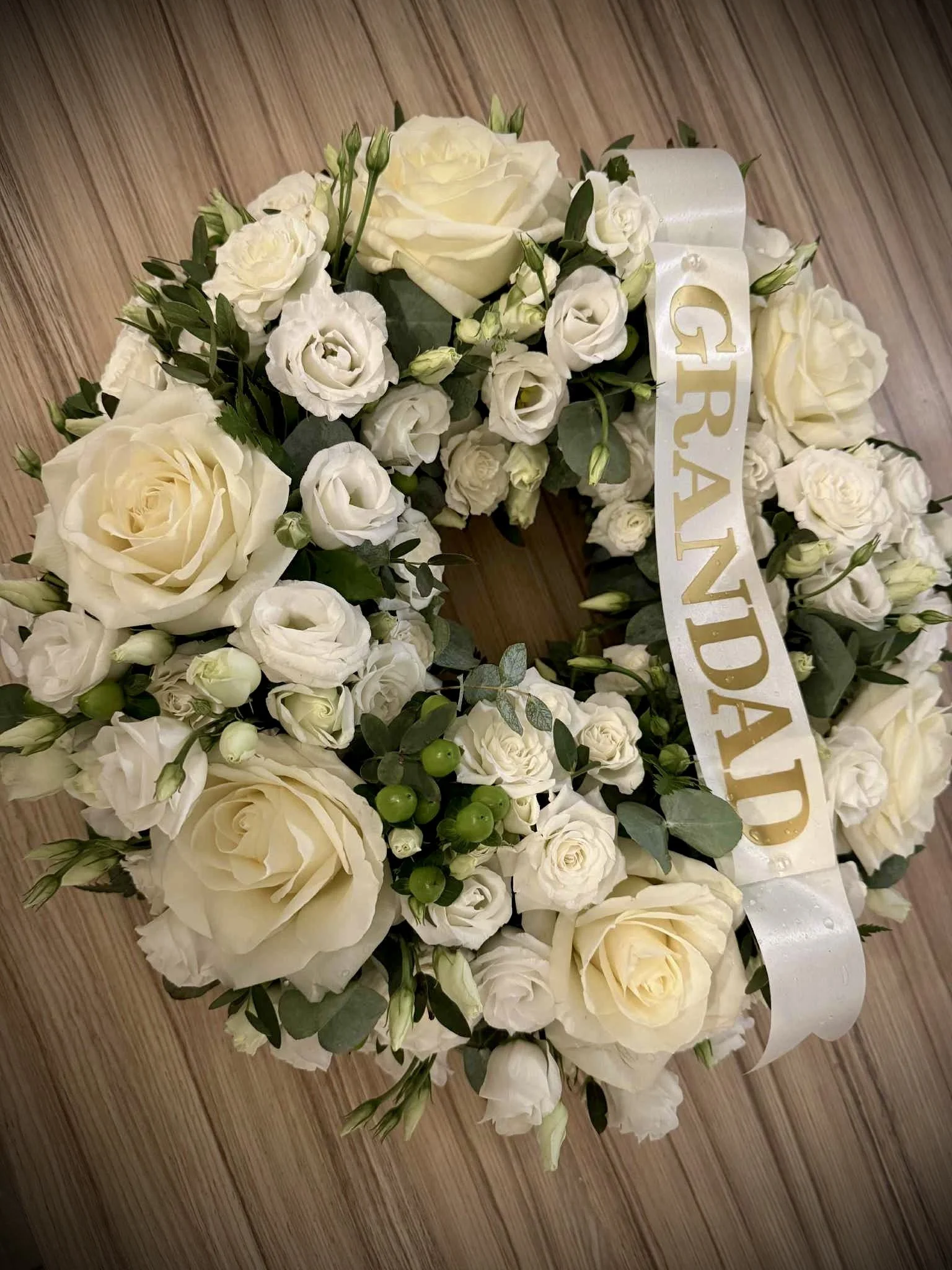 A floral arrangement of white roses, lisianthus, greenery, and a white satin ribbon with gold lettering reading 'GRAND'. The flowers are on a wooden surface.