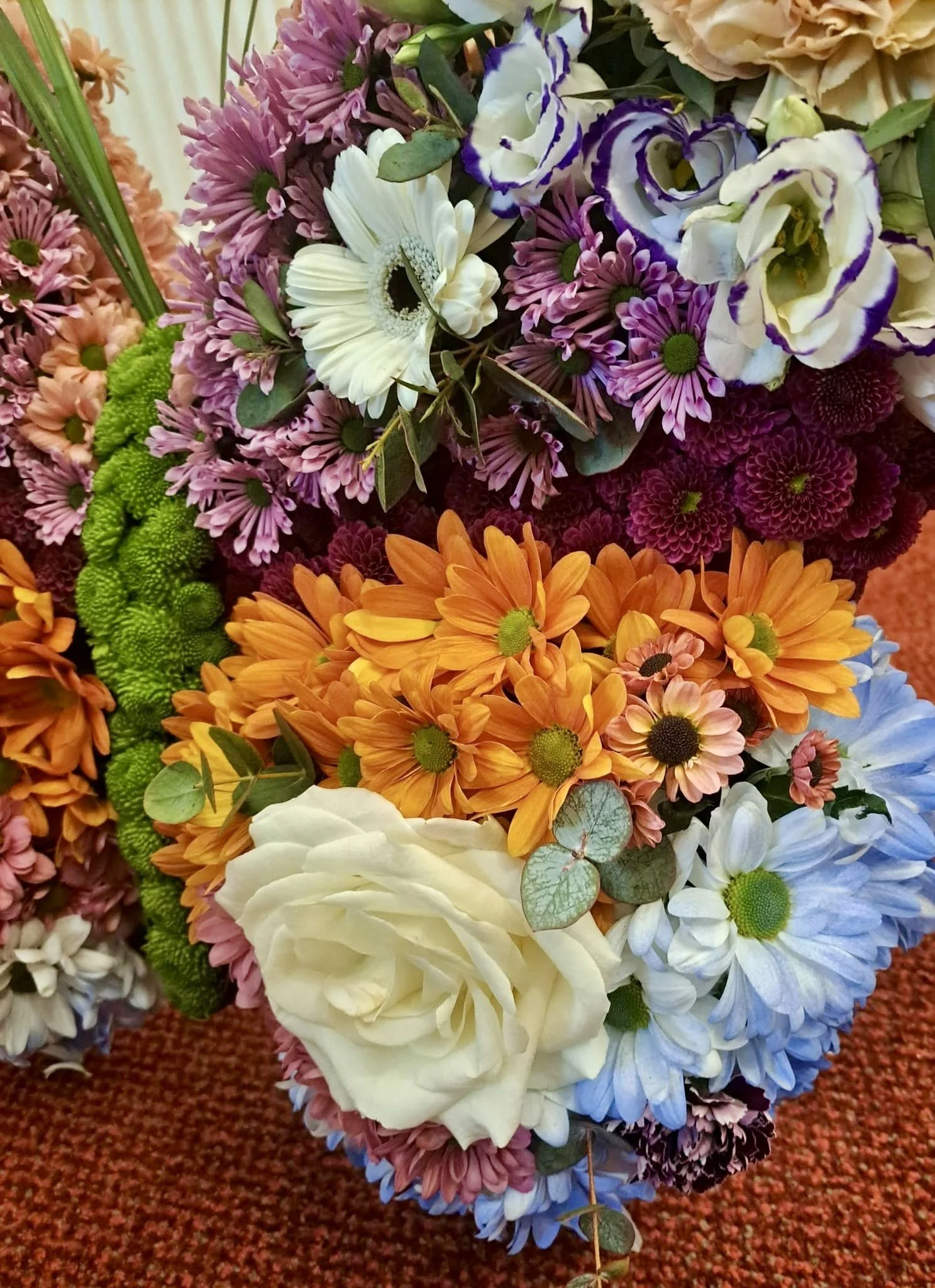 A bouquet of colorful flowers including purple, white, orange, and green blooms, arranged on a textured red surface.