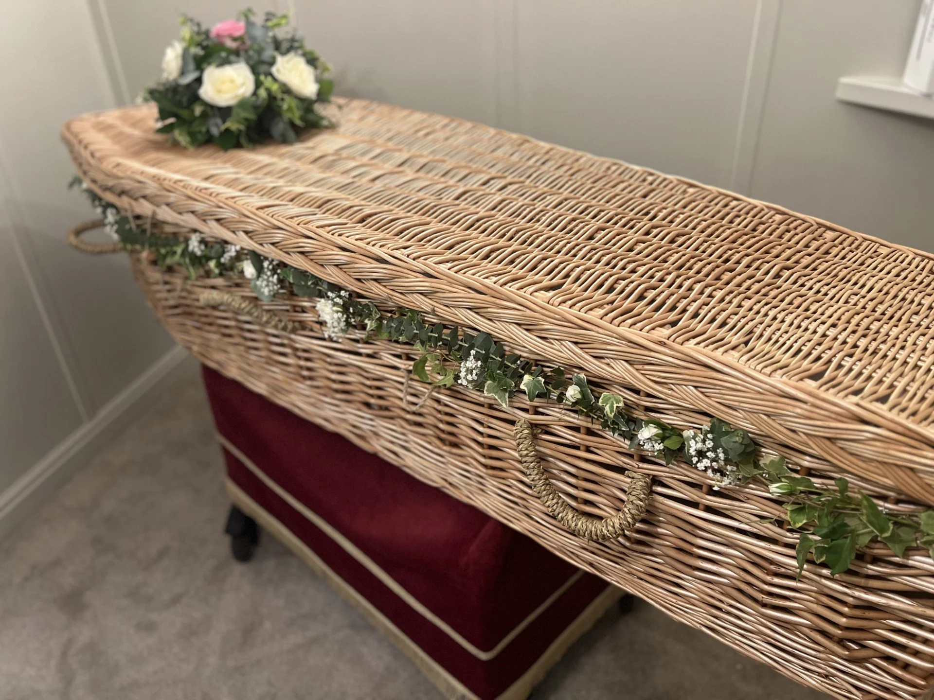 A wicker funeral casket decorated with flowers and green ivy rests on a red velvet stand inside a room with beige walls and carpeted floor.
