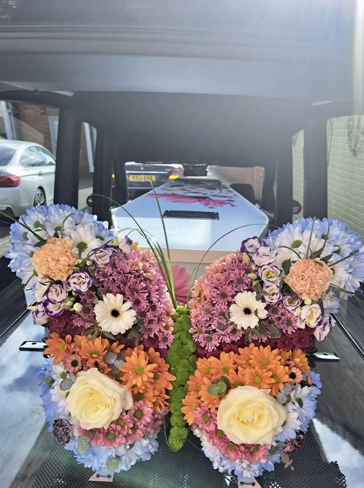 A funeral hearse decorated with a large arrangement of colorful flowers, including white, purple, pink, orange, and cream blooms, viewed from inside the vehicle looking out.