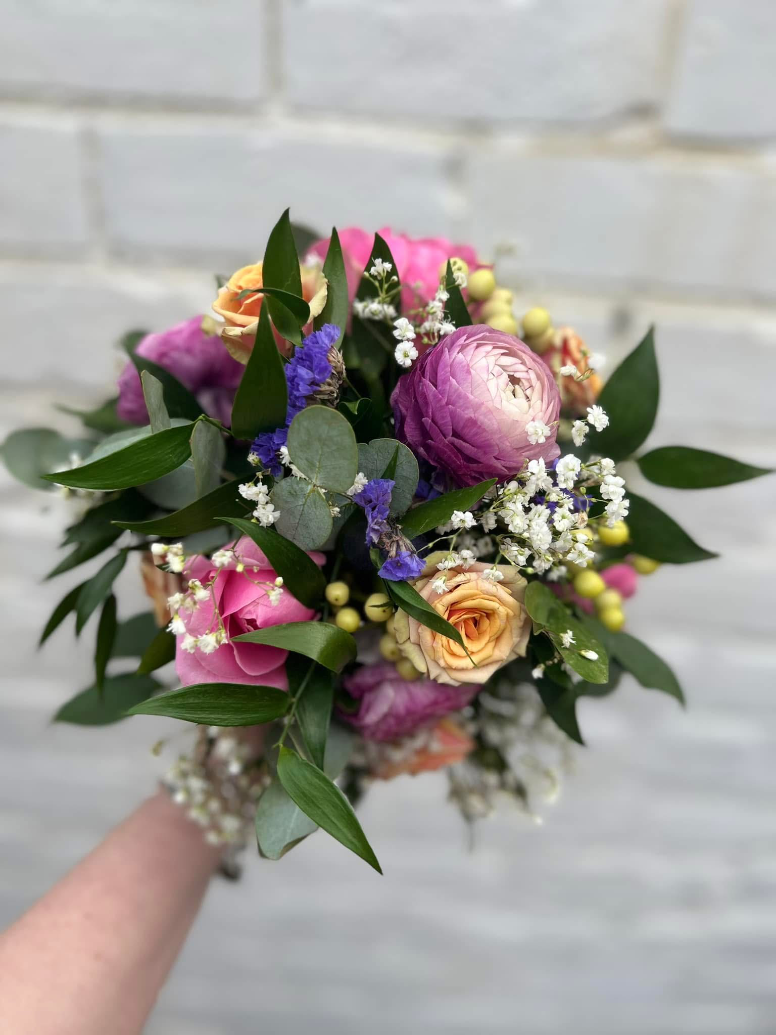 Hand holding a bouquet of pink, purple, orange, and white flowers with green leaves against a light gray background.