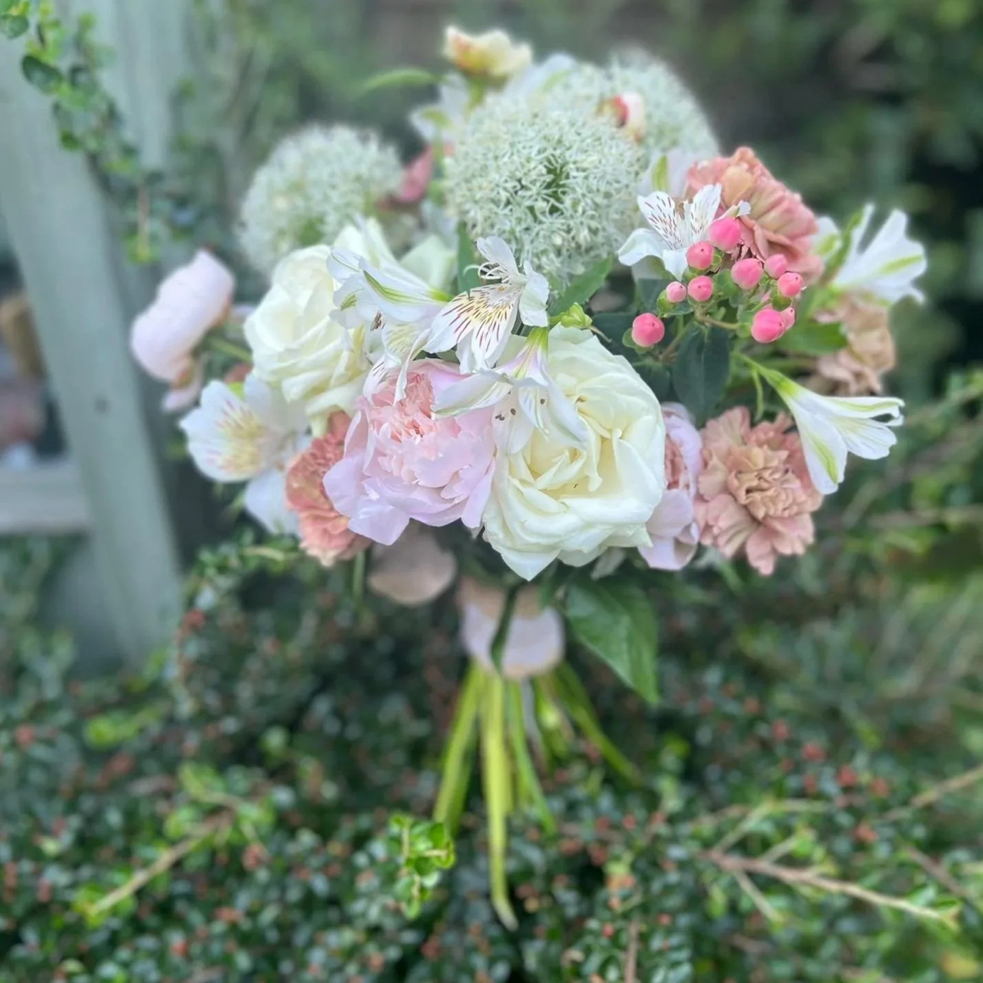 A bouquet of various light-colored flowers, including white roses, pink carnations, and small pink buds, held outdoors with a blurred green background.