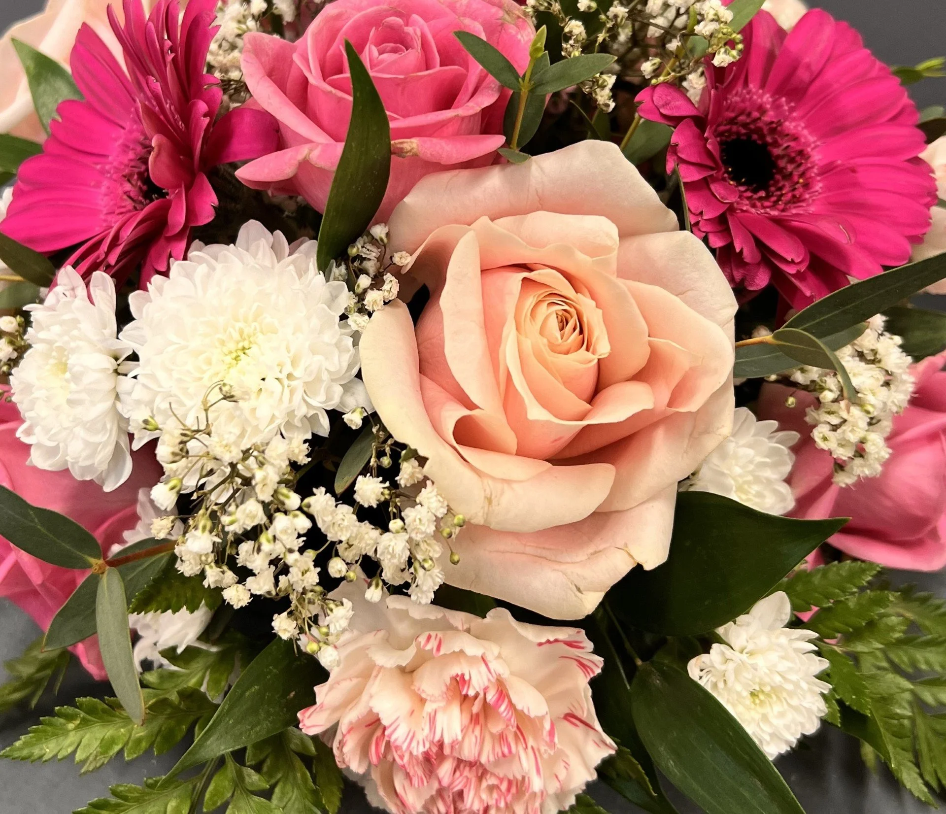 Close-up of a beautiful bouquet of mixed flowers including pink roses, white chrysanthemums, pink gerbera daisies, and small white baby's breath, accented with green leaves.
