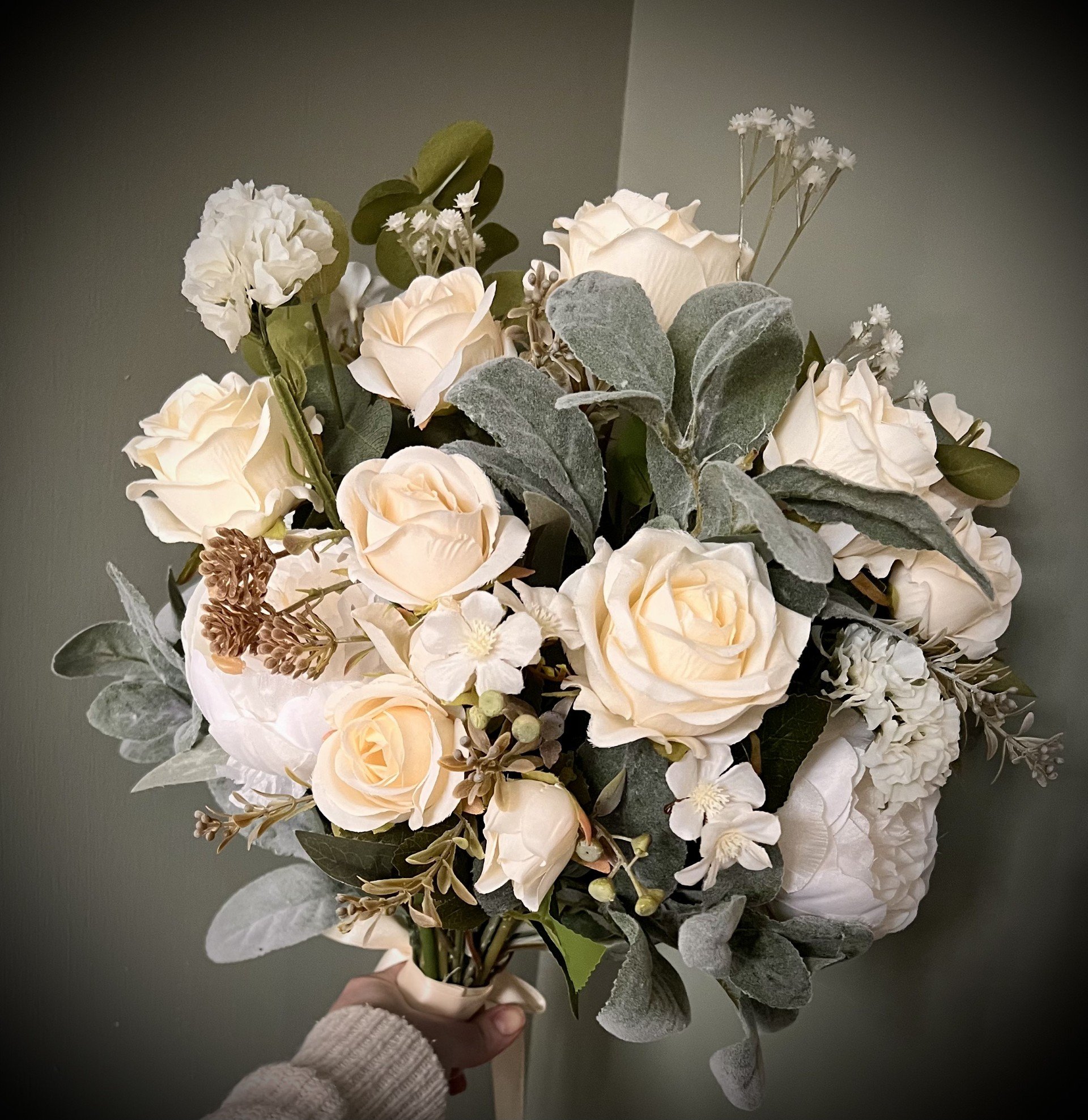 A bouquet of cream-colored roses, white hydrangeas, and small white filler flowers held by a hand wearing a white knitted sweater against a gray background.